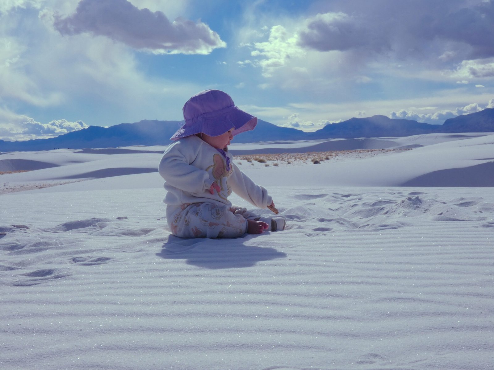 A baby sitting and playing in the bright white gypsum sand with the San Andres Mountains in the background at White Sands National Park.