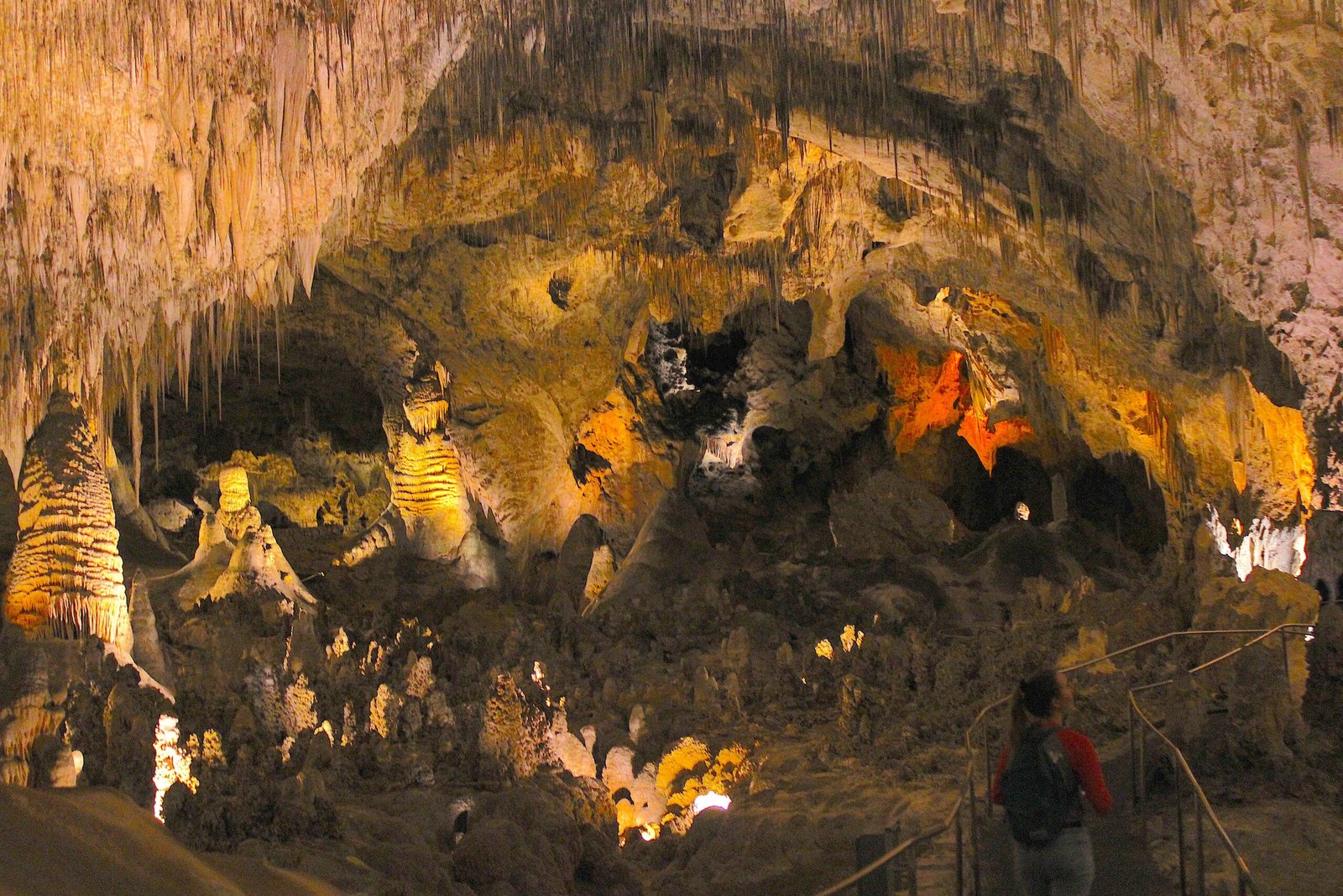 Woman walking along a handrail path inside Carlsbad Caverns gazing up at massive stalactites and cave formations.