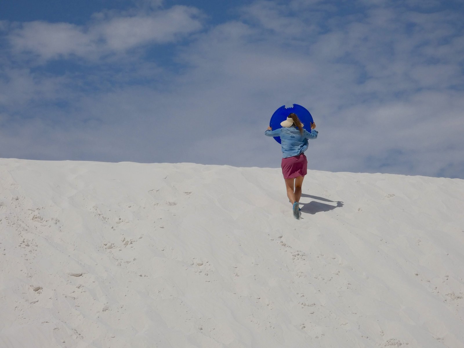 A woman carrying a plastic saucer sled over her head while walking up a steep white gypsum sand dune at White Sands National Park.