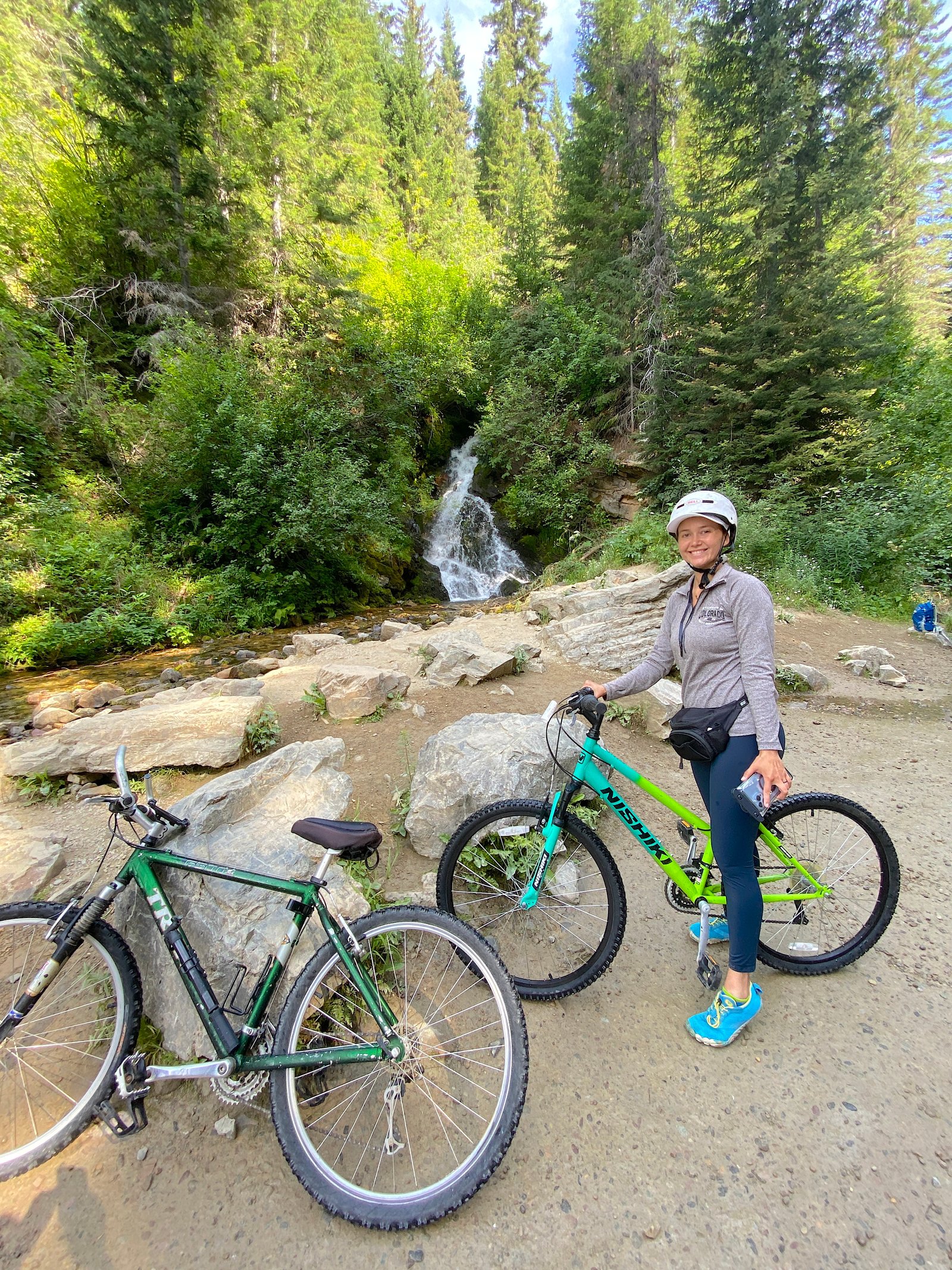 Smiling female cyclist with two bicycles standing in front of a lush mountain waterfall surrounded by green trees after emerging from the Taft Tunnel on the Route of the Hiawatha.