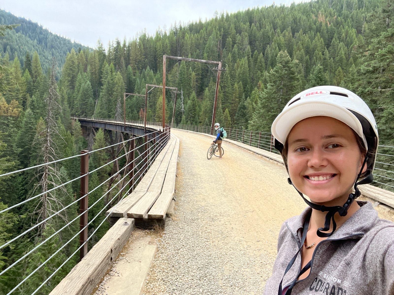 Selfie of a smiling woman on a bicycle with her husband riding his bike on a wooden trestle bridge in the background on the Route of the Hiawatha.