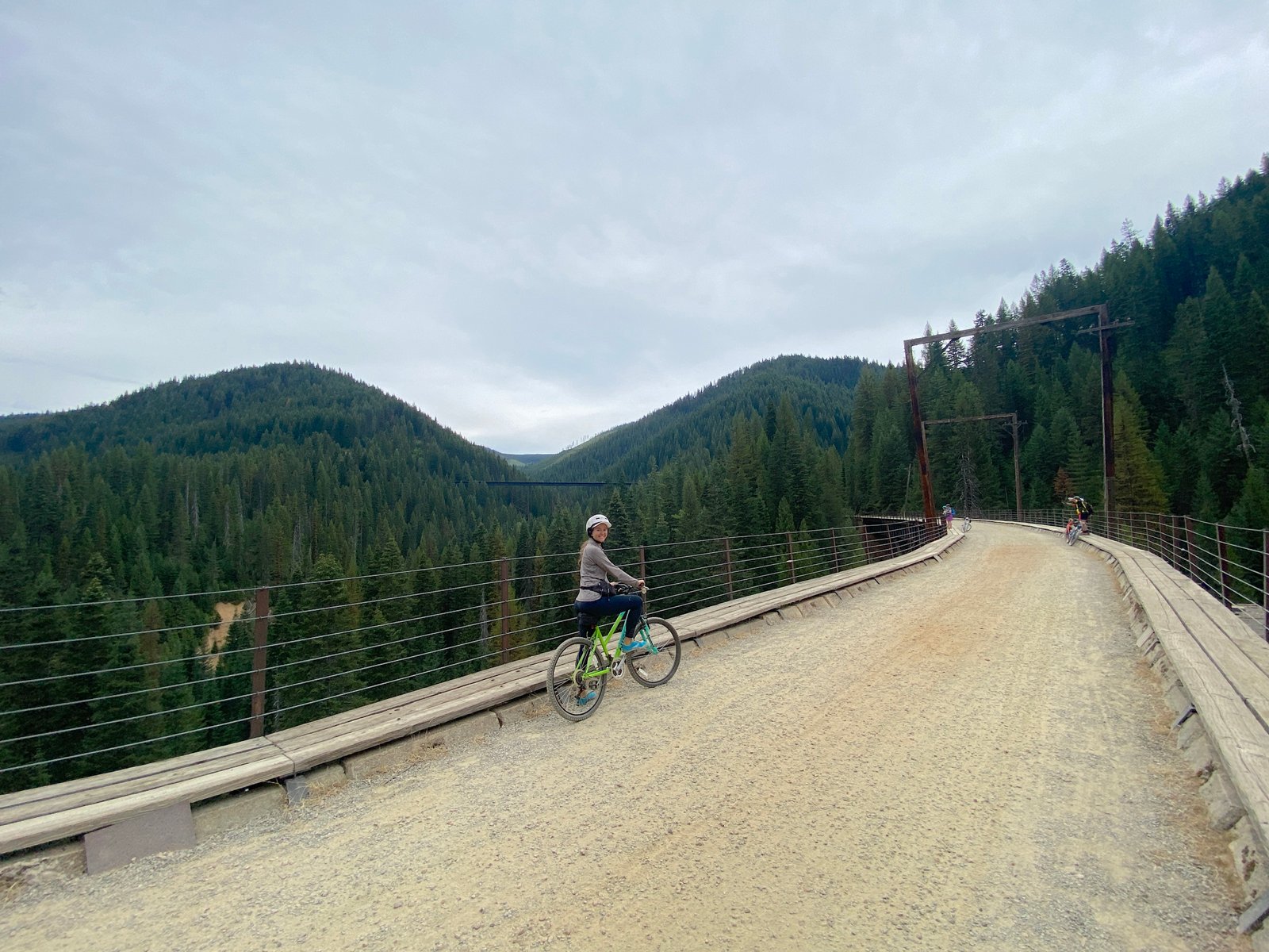 Smiling female cyclist with her bicycle standing on the wooden deck of a high trestle bridge with dense forested mountains in the background on the Route of the Hiawatha.