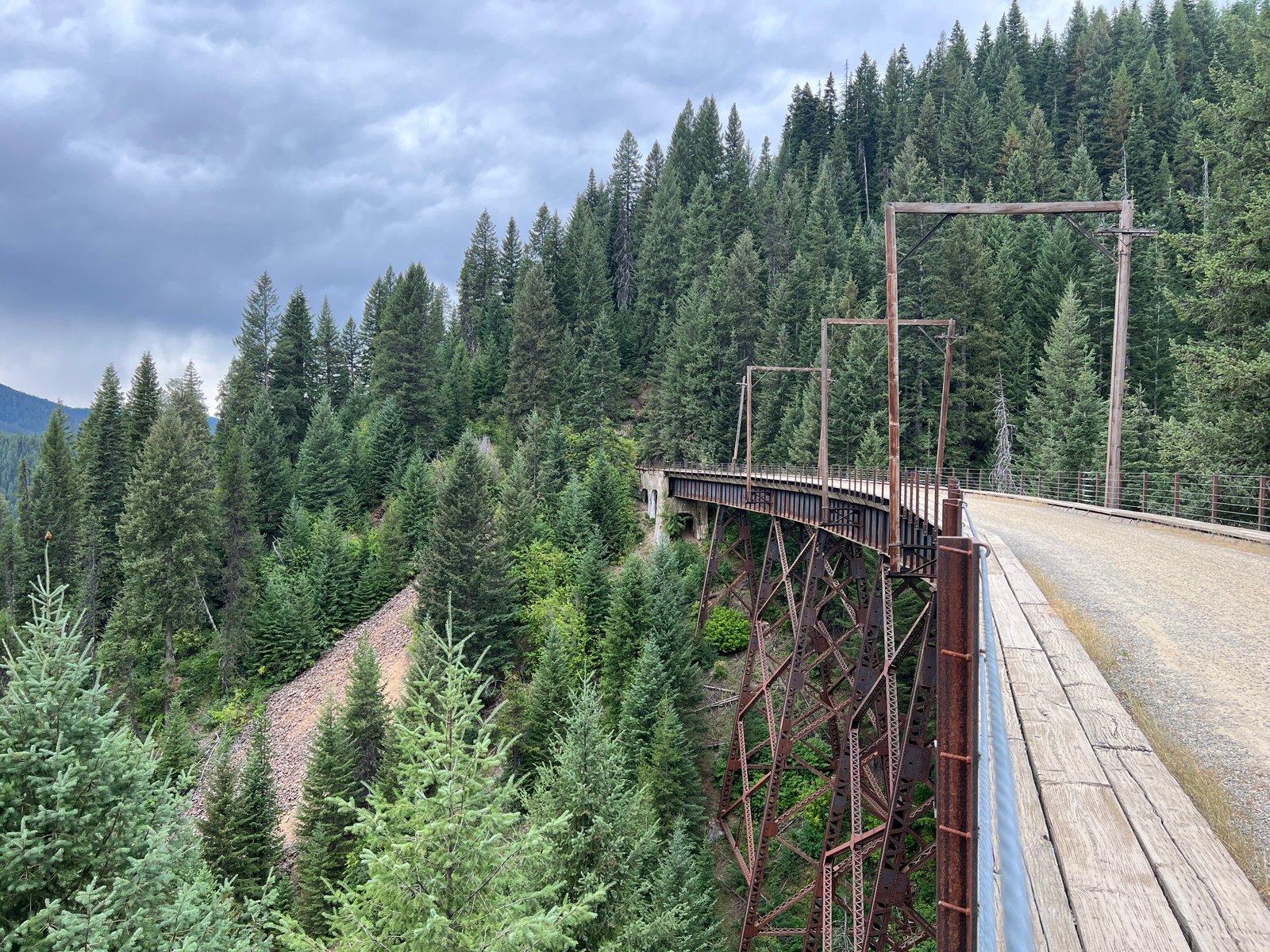 Side angle view of a tall wooden trestle train bridge spanning a dense pine tree mountain under dark, stormy summer clouds on the Route of the Hiawatha.