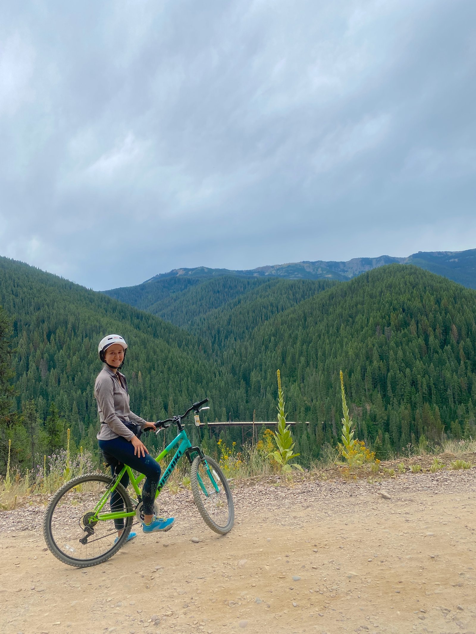 Smiling female cyclist sitting on her bicycle along a dirt rail-trail with dense, green forested mountains in the background on the Route of the Hiawatha.
