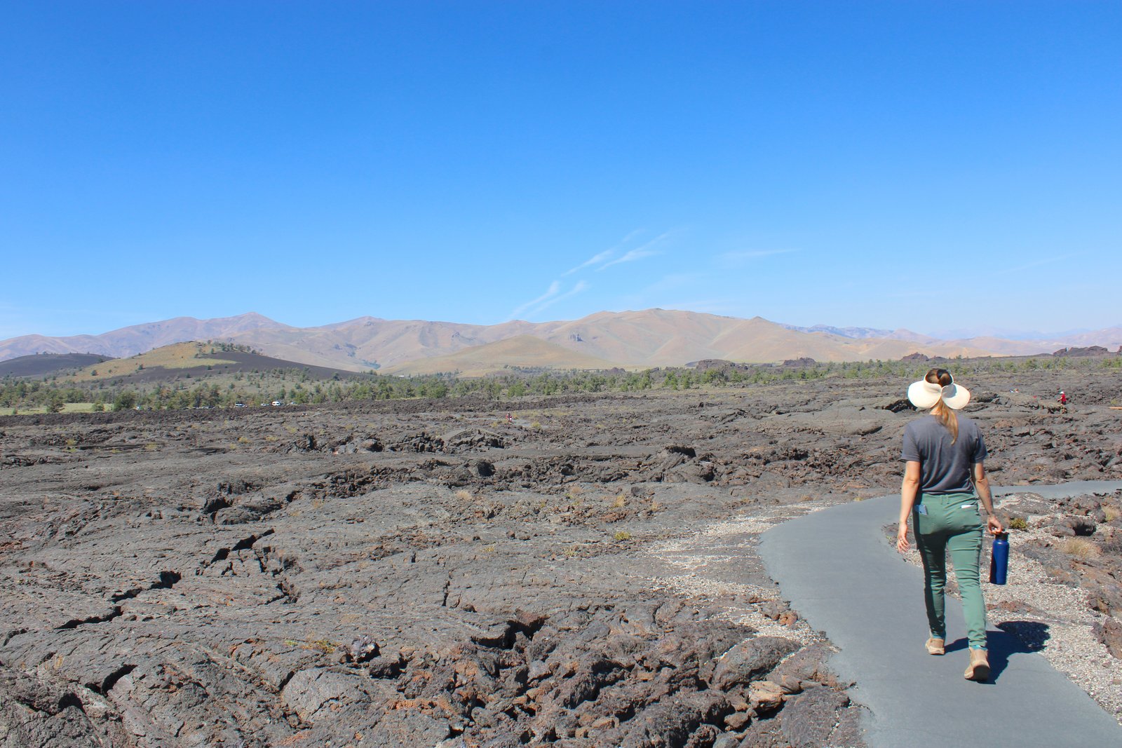 Hiker wearing a wide-brim sun hat and hiking boots holding a water bottle on a paved trail at Craters of the Moon