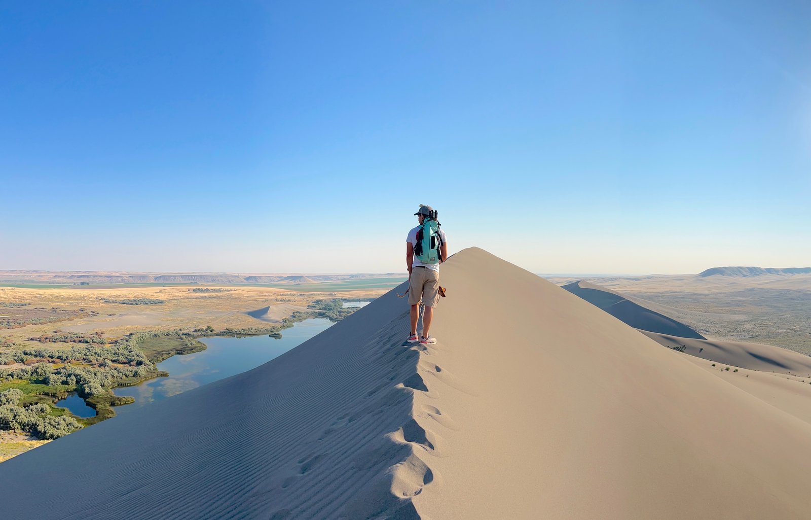 A hiker standing on the crest of a massive sand dune overlooking the desert lakes and distant mountains at Bruneau Dunes State Park in Idaho.