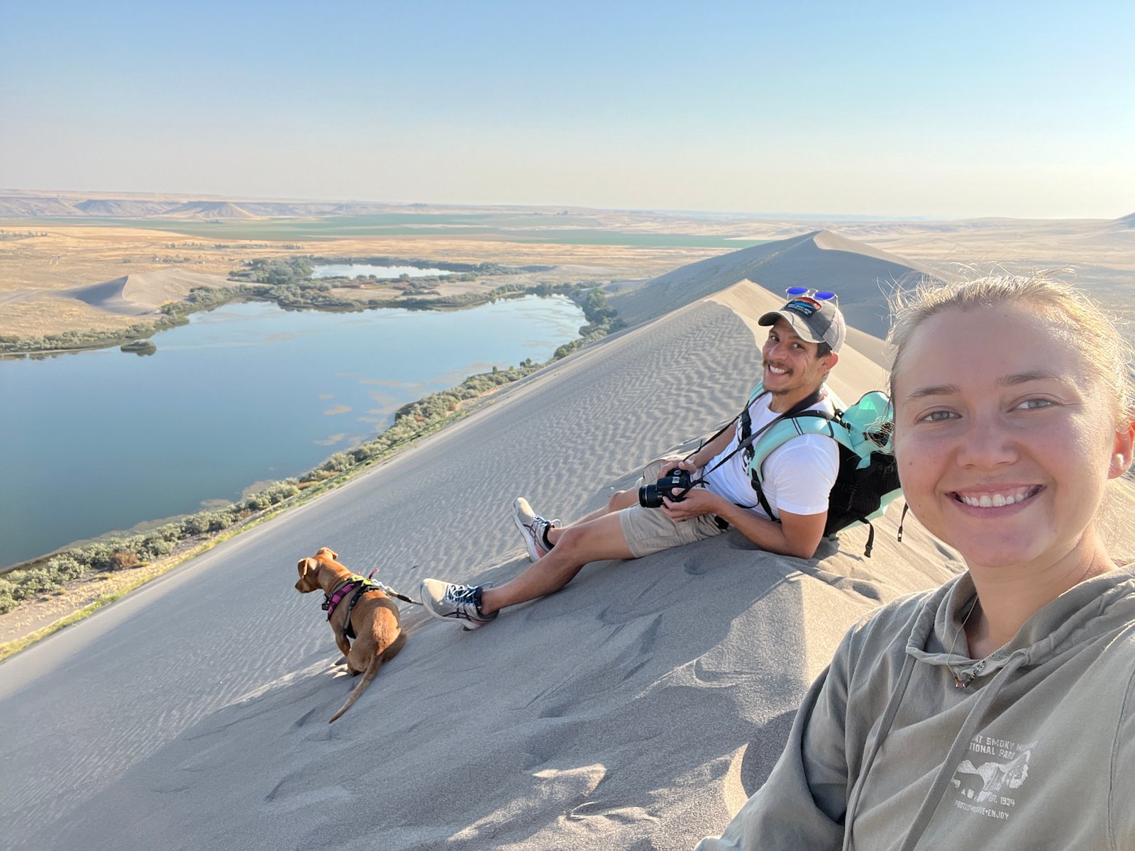 A couple and their dog taking a selfie while sitting on the crest of the sand dunes at Bruneau Dunes State Park, overlooking the lakes and mountains at sunset.