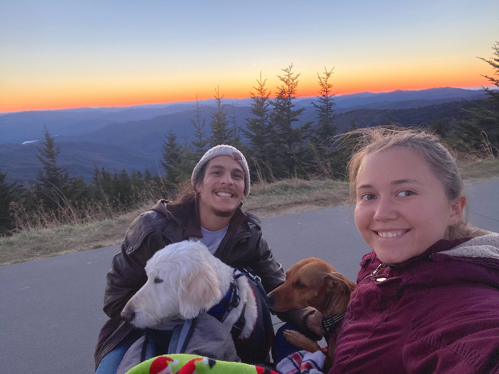 A selfie of a couple smiling with their two dogs outdoors against a vibrant mountain sunset background.