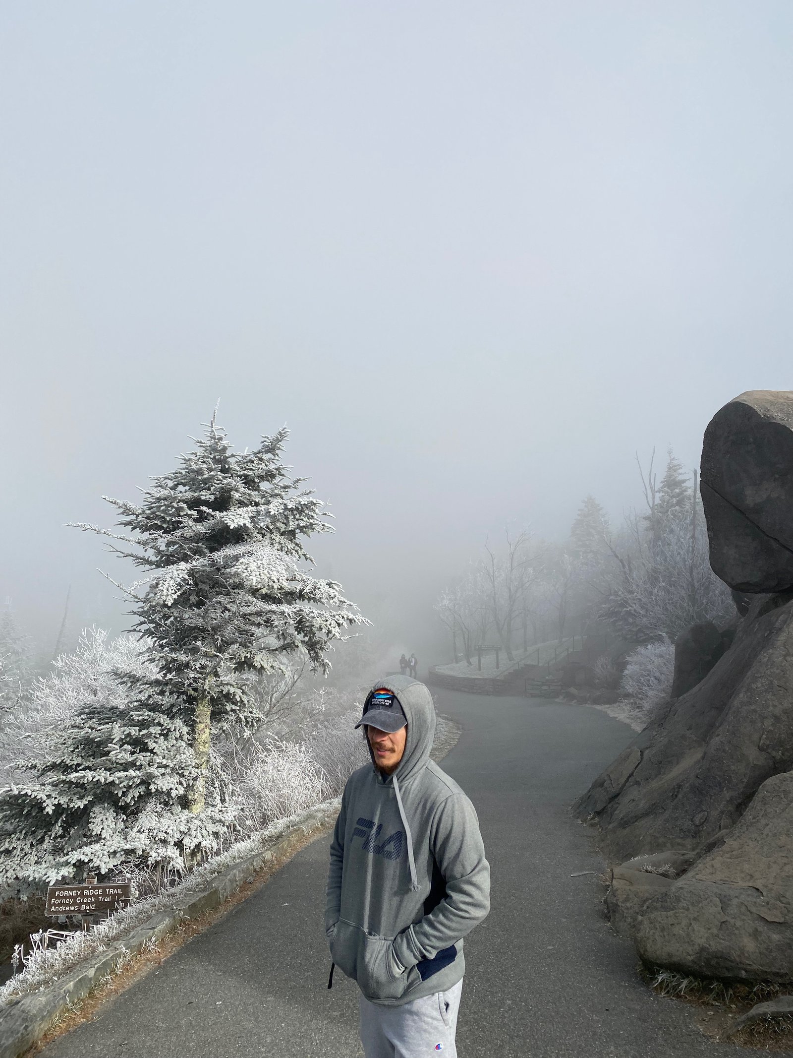 A person in a hoodie standing on a misty, fog-shrouded mountain overlook with frosted pine trees and rocky terrain in the Great Smoky Mountains National Park.