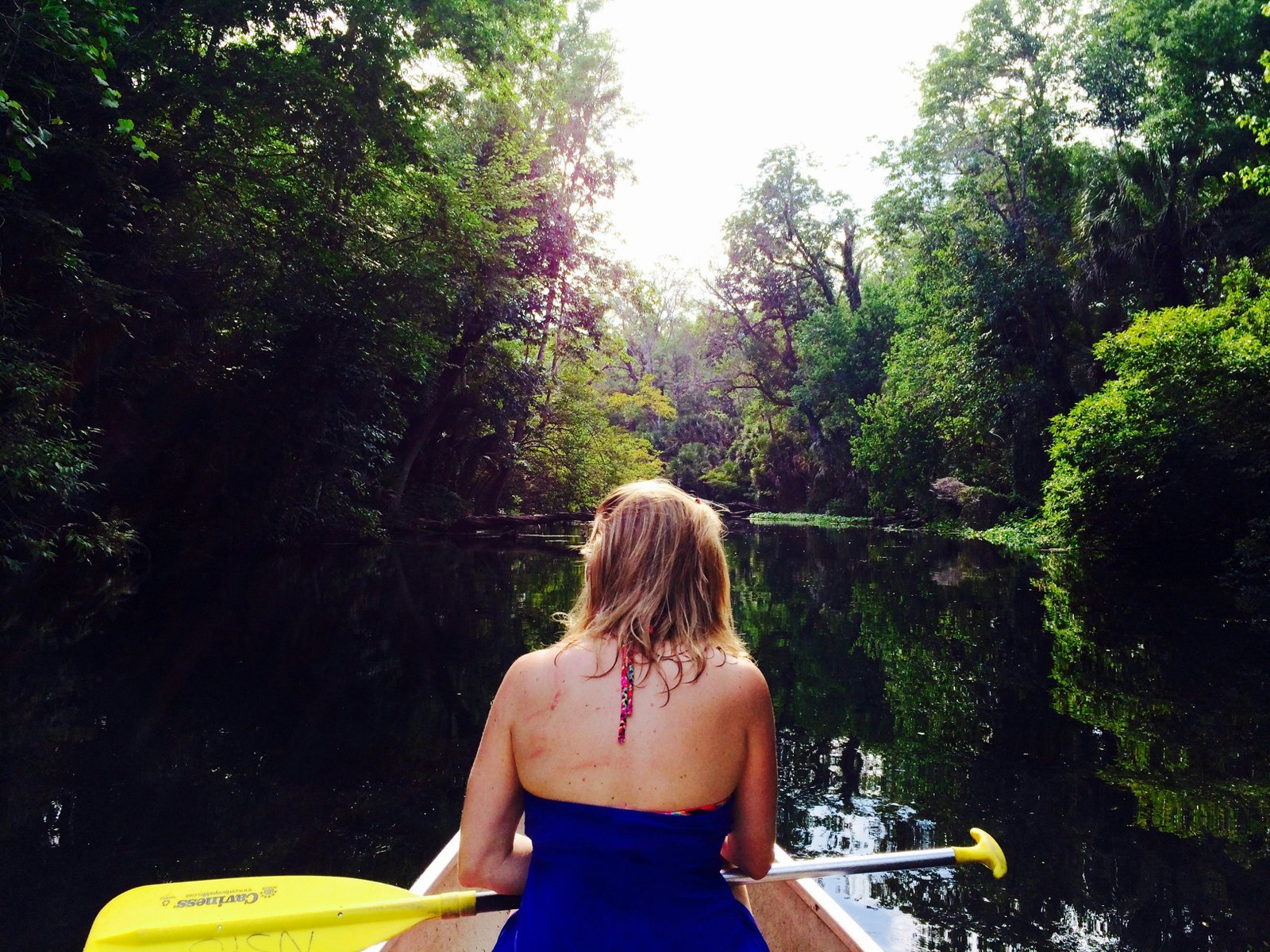 A person paddling a canoe or kayak down a calm, tree-lined river run reflecting lush green forest foliage at Wekiwa Springs State Park.