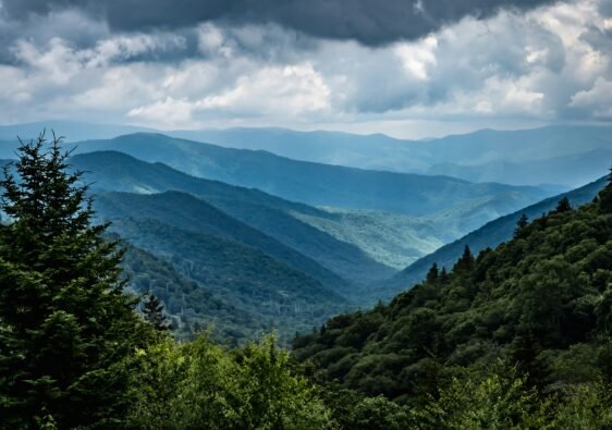 A sweeping panoramic view of green rolling mountain ridges fading into a cloudy blue sky from a high-elevation overlook in the Smokies.