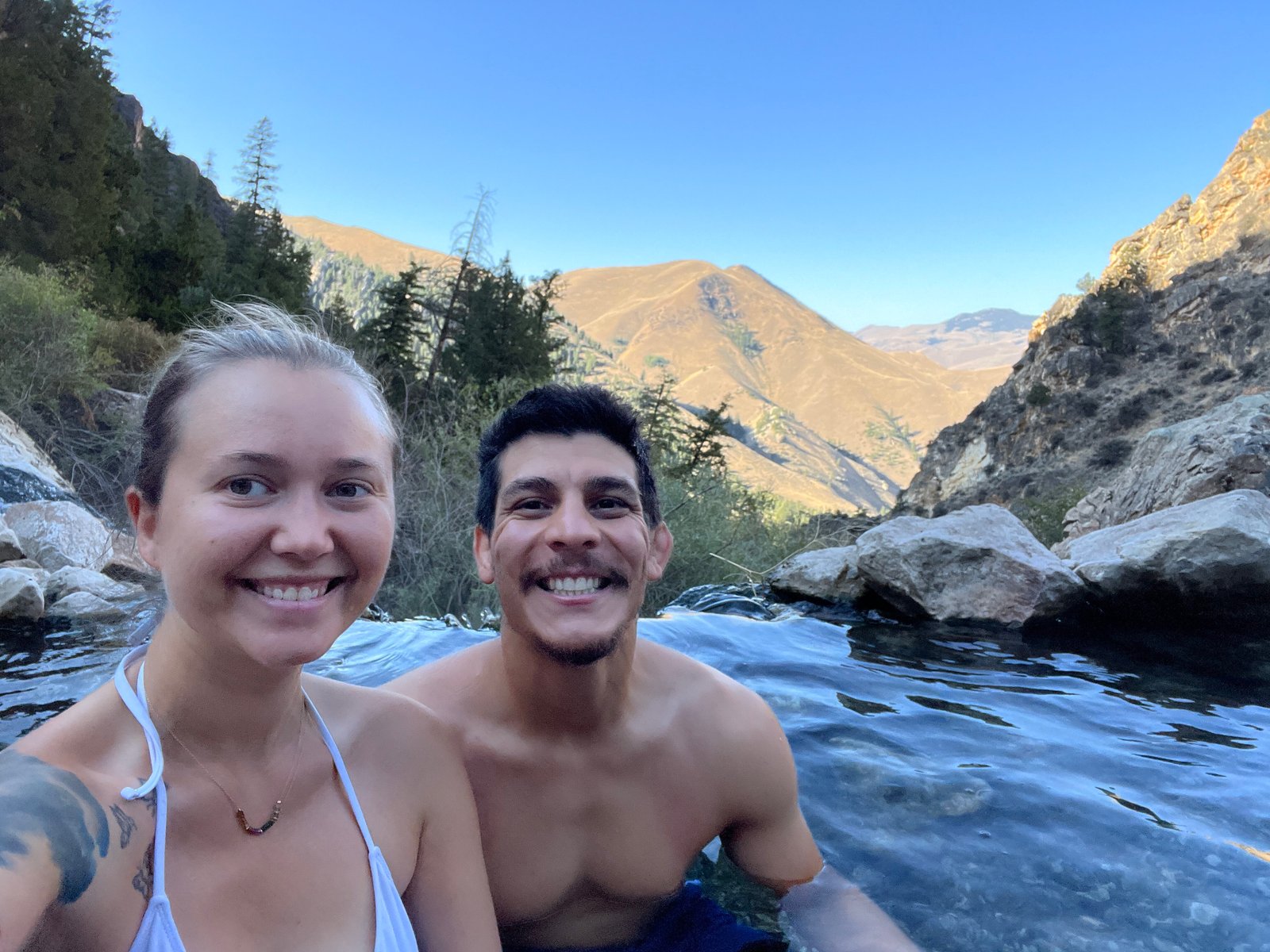 A smiling couple taking a selfie while soaking in a thermal pool at Goldbug Hot Springs with mountain peaks behind them.