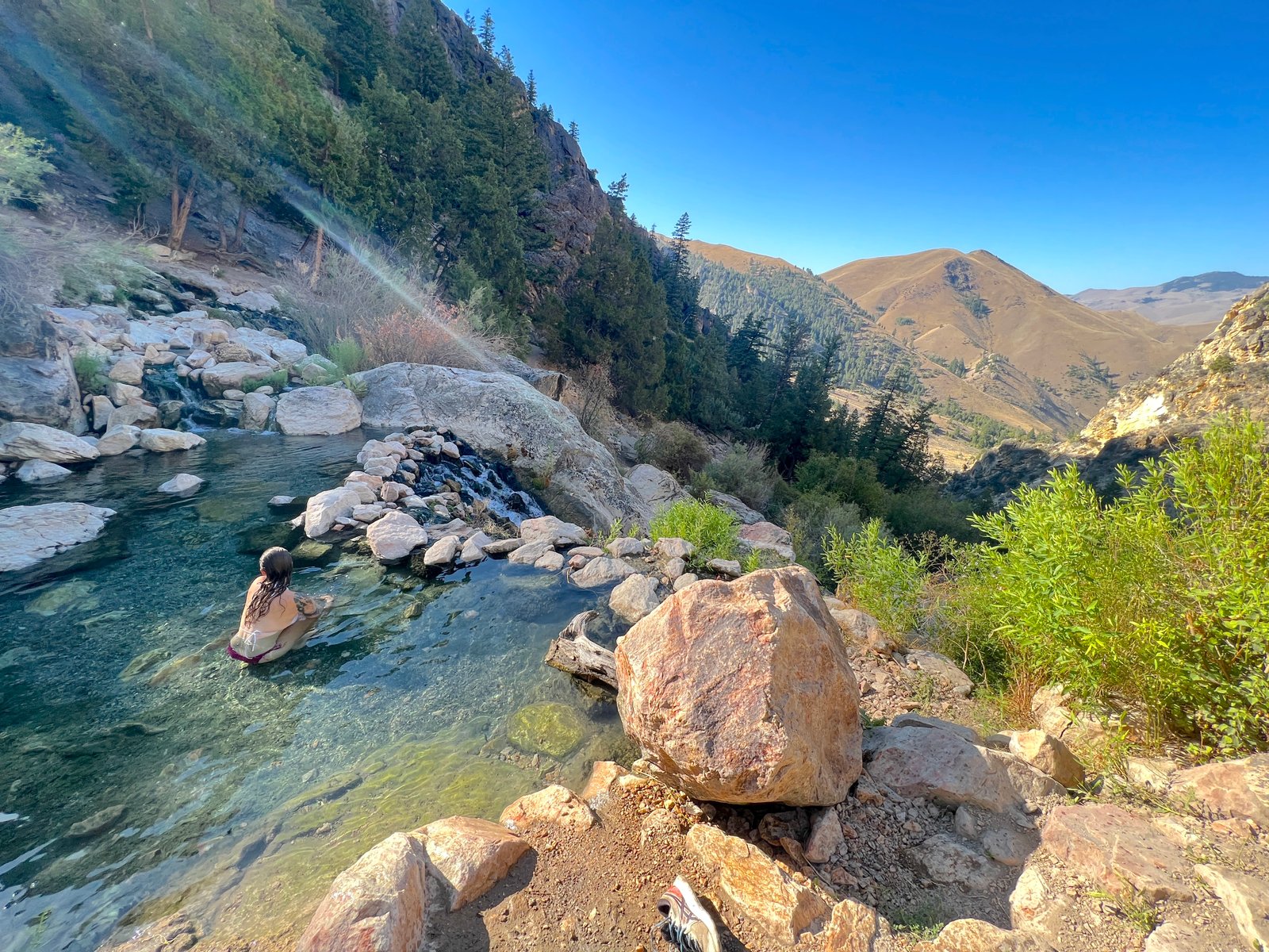 A woman sitting in a natural thermal pool at Goldbug Hot Springs, looking out over a scenic mountain valley.