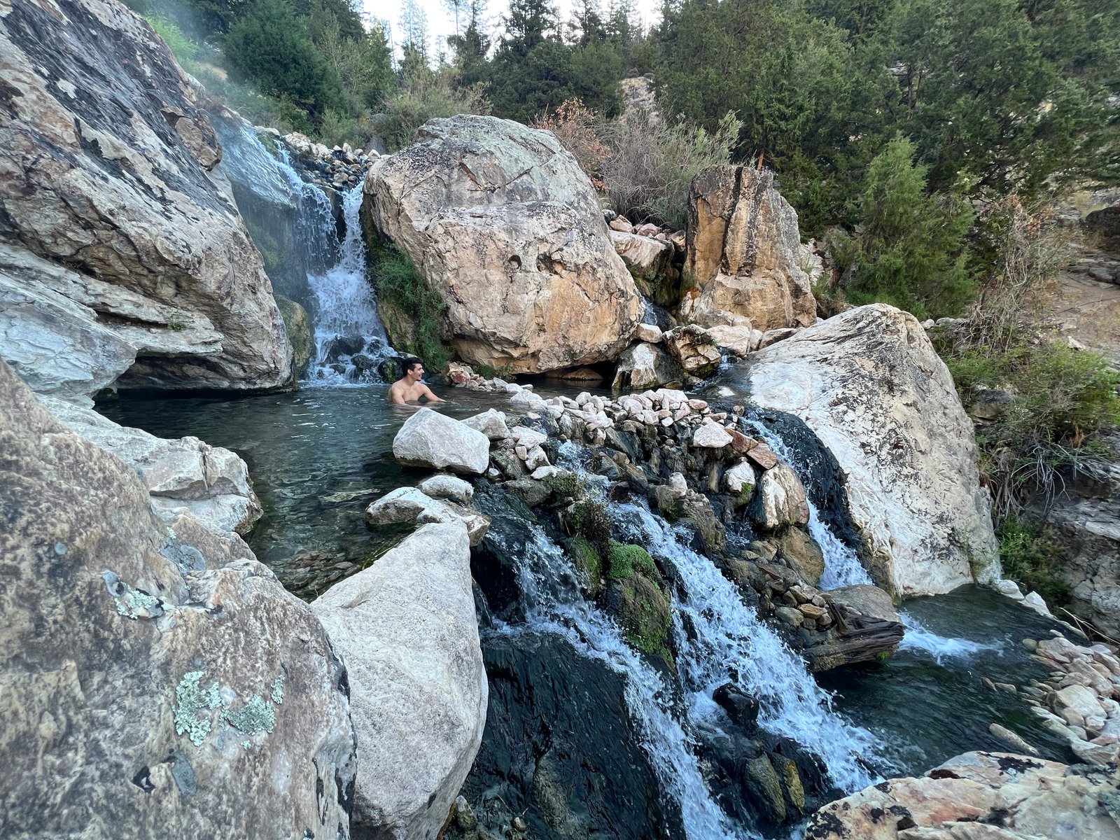 Wide landscape view of a man soaking in a natural rock pool at Goldbug Hot Springs surrounded by mountains and desert valley scenery.
