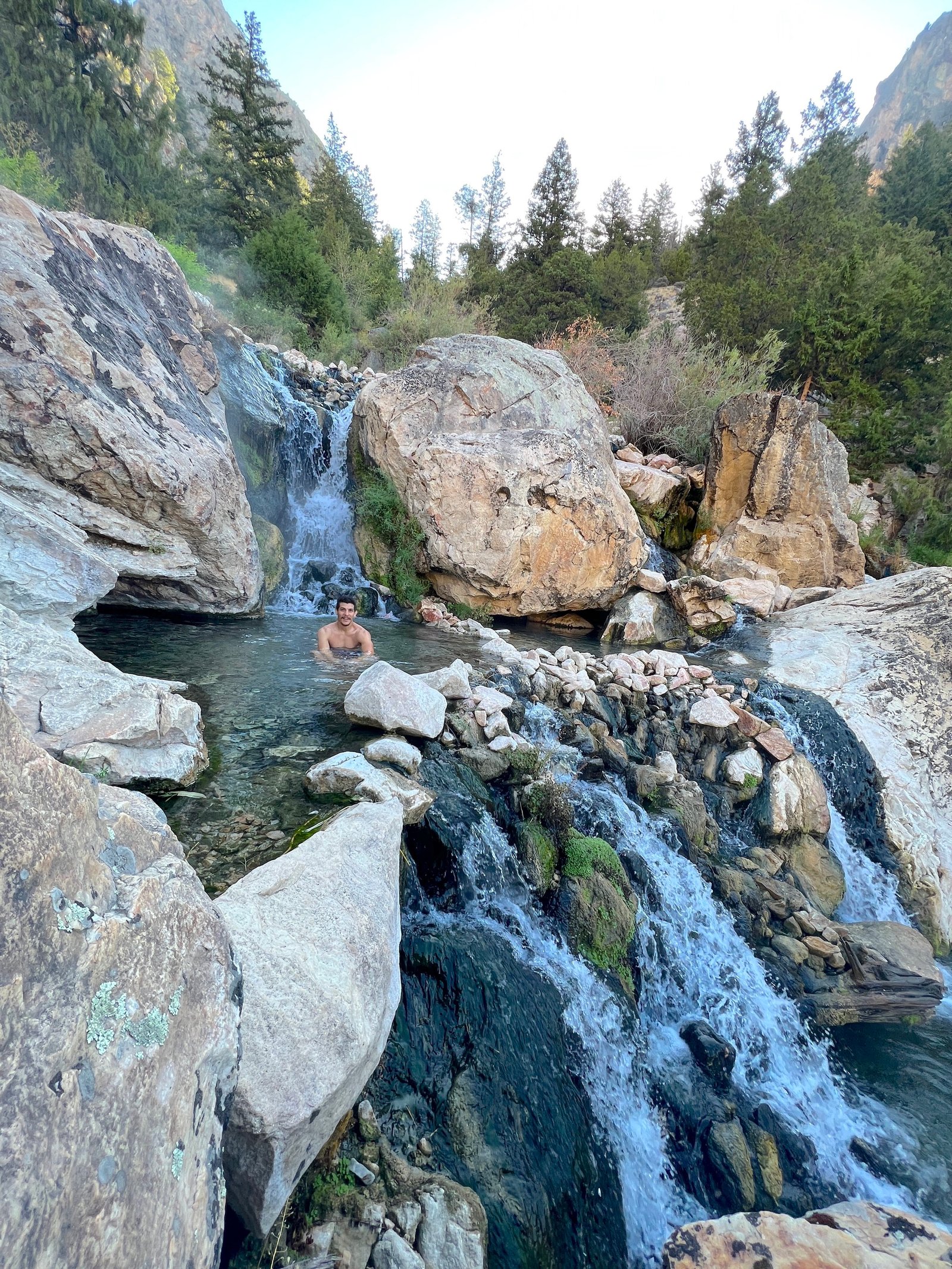 A man relaxing in a natural thermal pool at Goldbug Hot Springs as cascading waterfalls pour into the lower pools behind him.