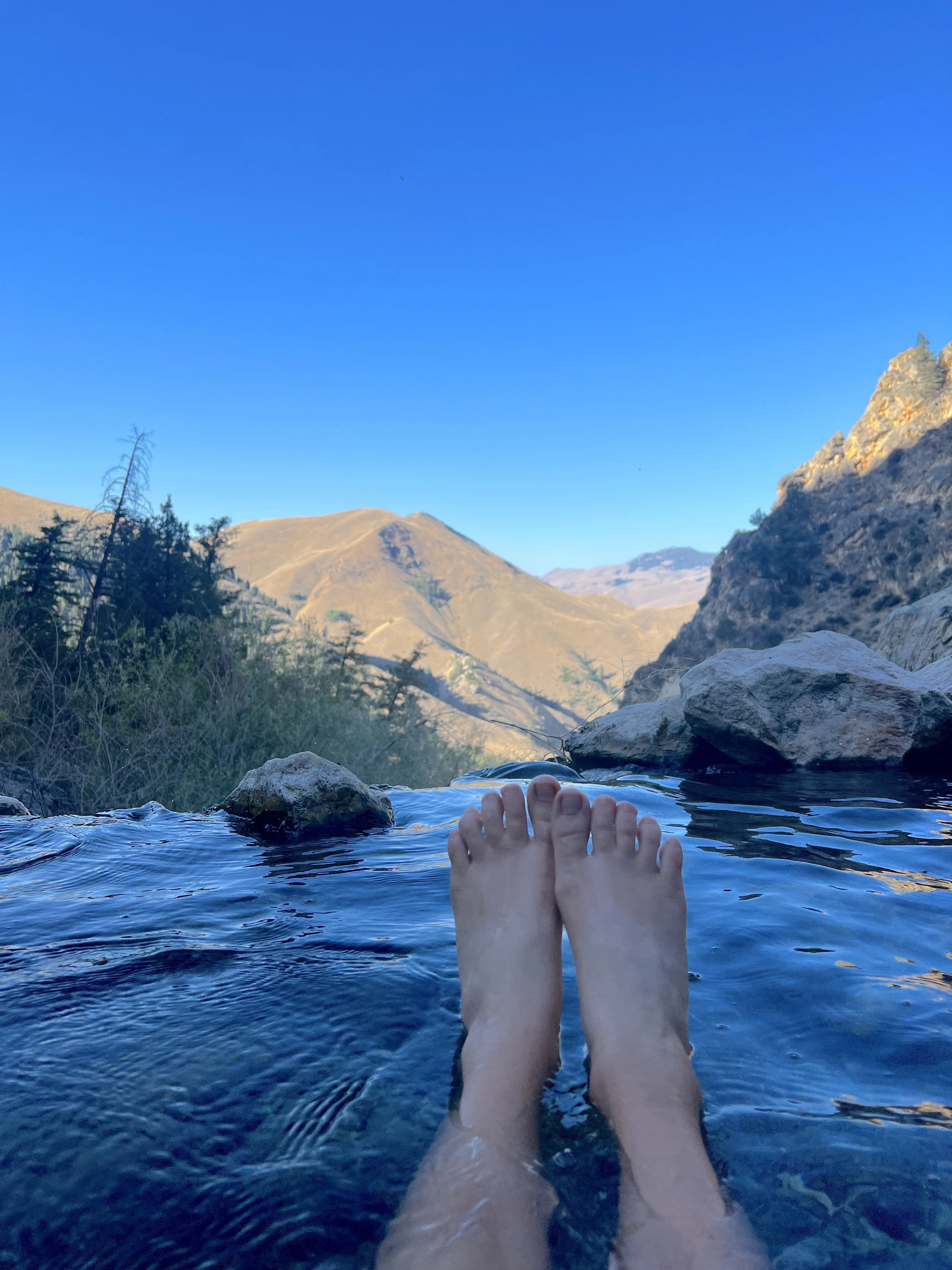 First-person point-of-view shot of a person's feet sticking out of warm water at Goldbug Hot Springs, with snow-capped mountains in the background.