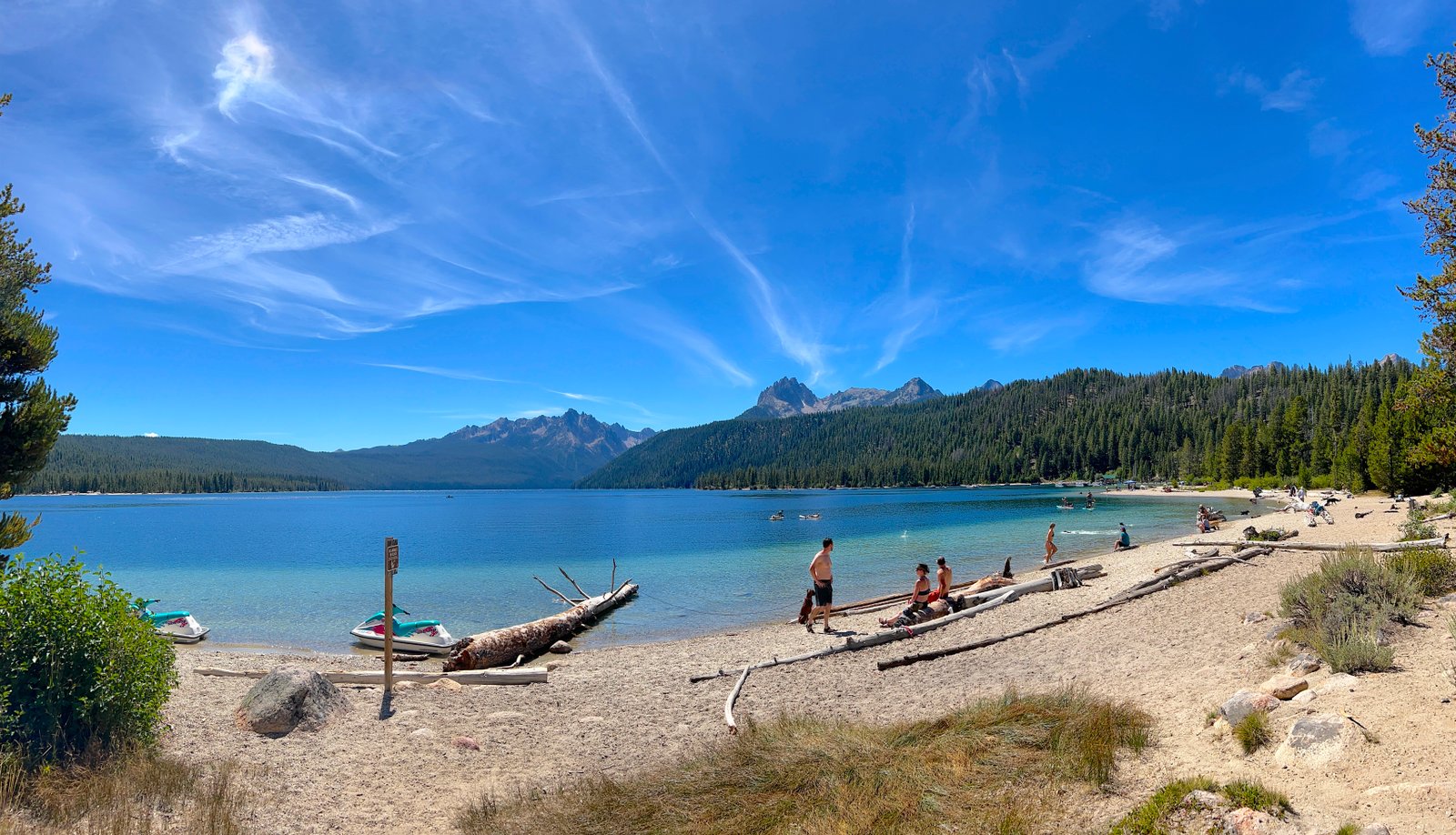 Visitors relaxing along the vibrant blue shoreline of Redfish Lake with pine trees and the jagged peaks of the Sawtooth Mountains in the distance, Idaho.