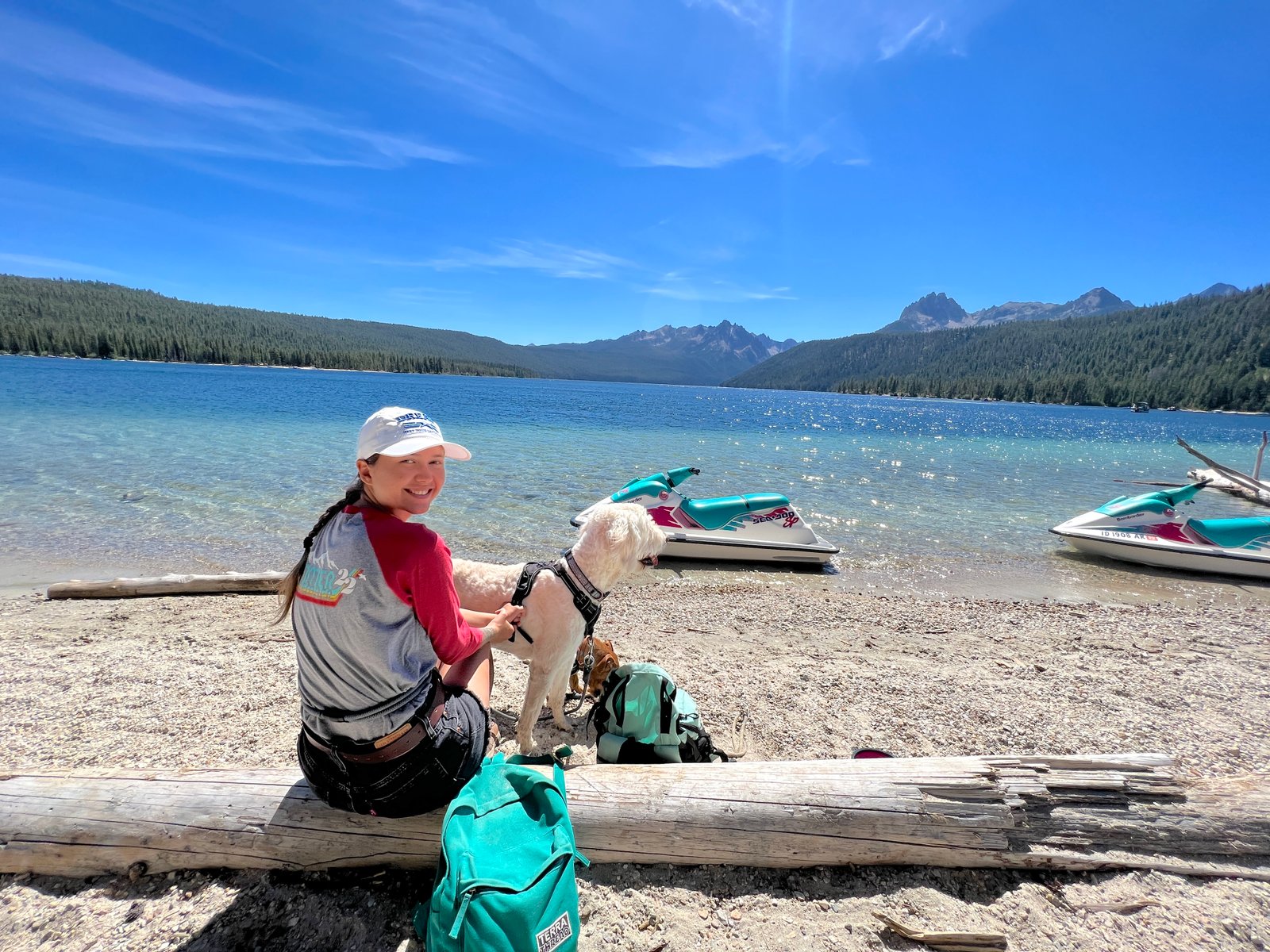 A visitor relaxing with a dog along the crystal-clear shoreline of Redfish Lake with jet skis and the Sawtooth Mountains in the background, Idaho.