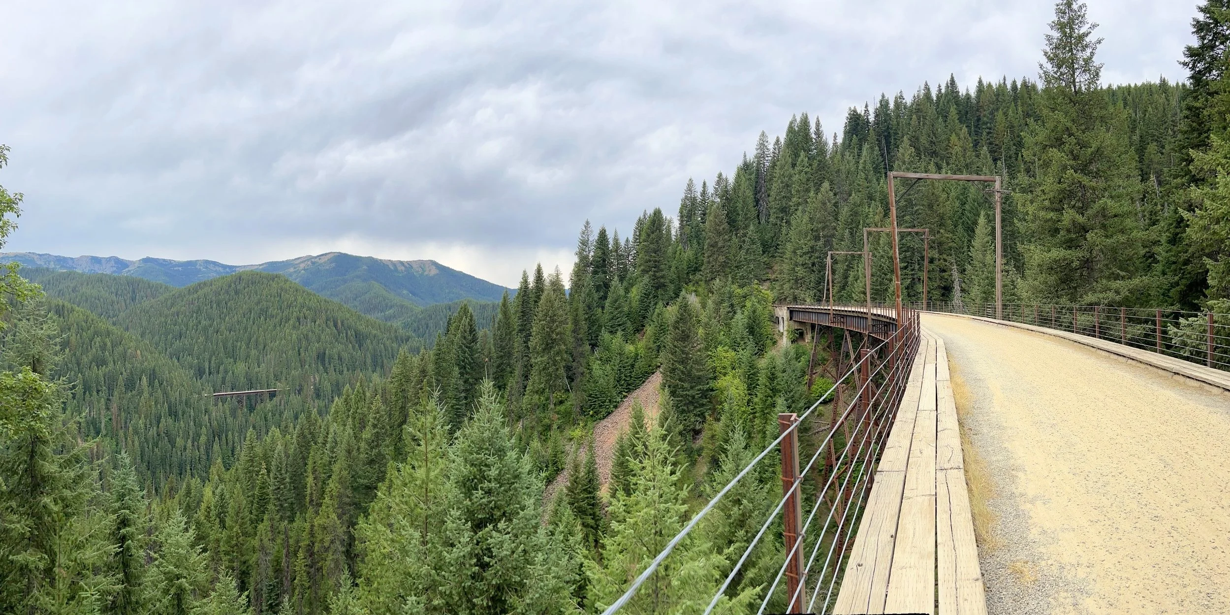Wide-angle view of a wooden trestle bridge on the Route of the Hiawatha bike trail, surrounded by dense forested mountains in Idaho with another bridge visible in the distance.