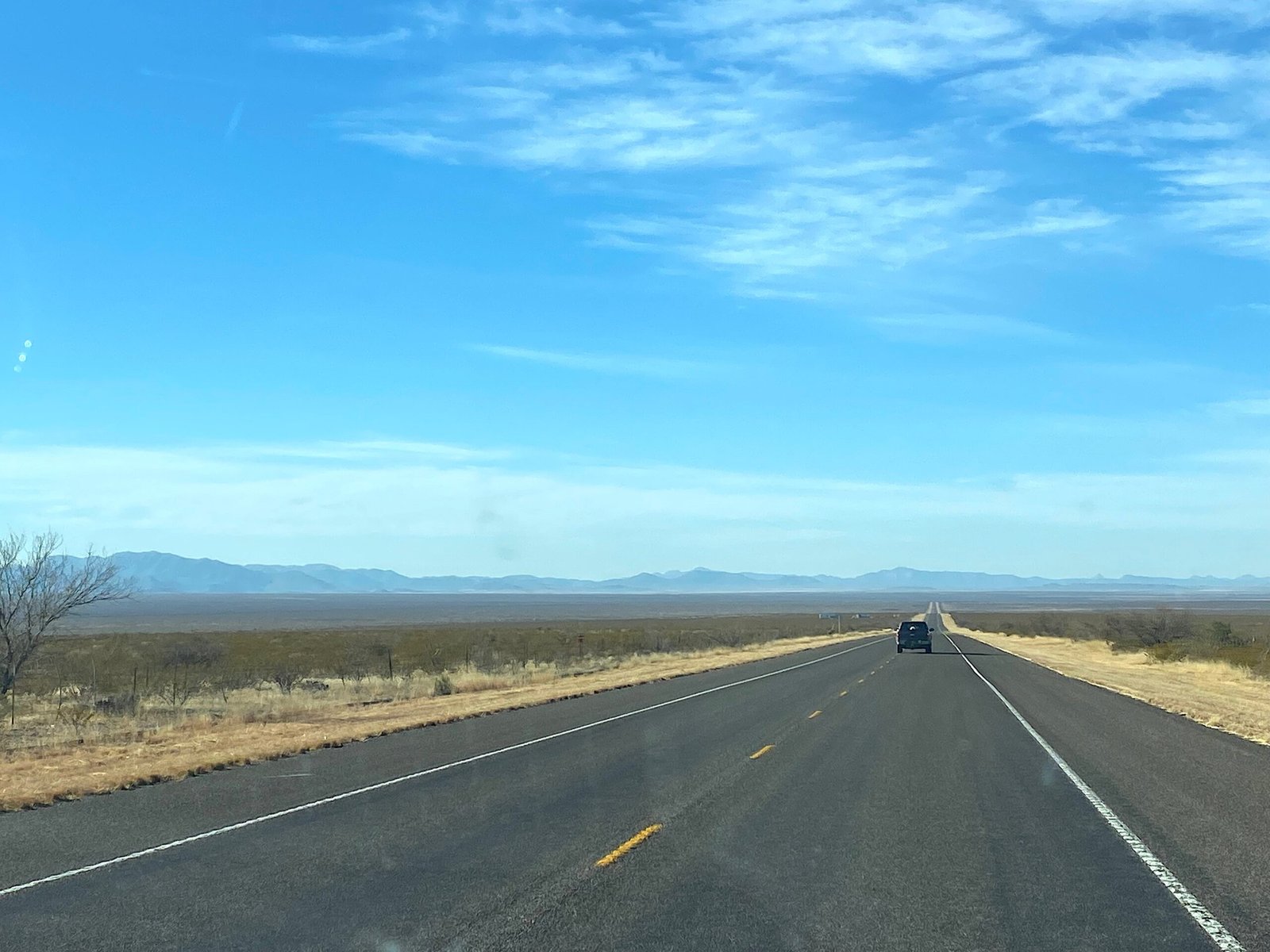 Long, straight two-lane highway stretching across the flat West Texas desert landscape toward distant mountain ranges under a blue sky.