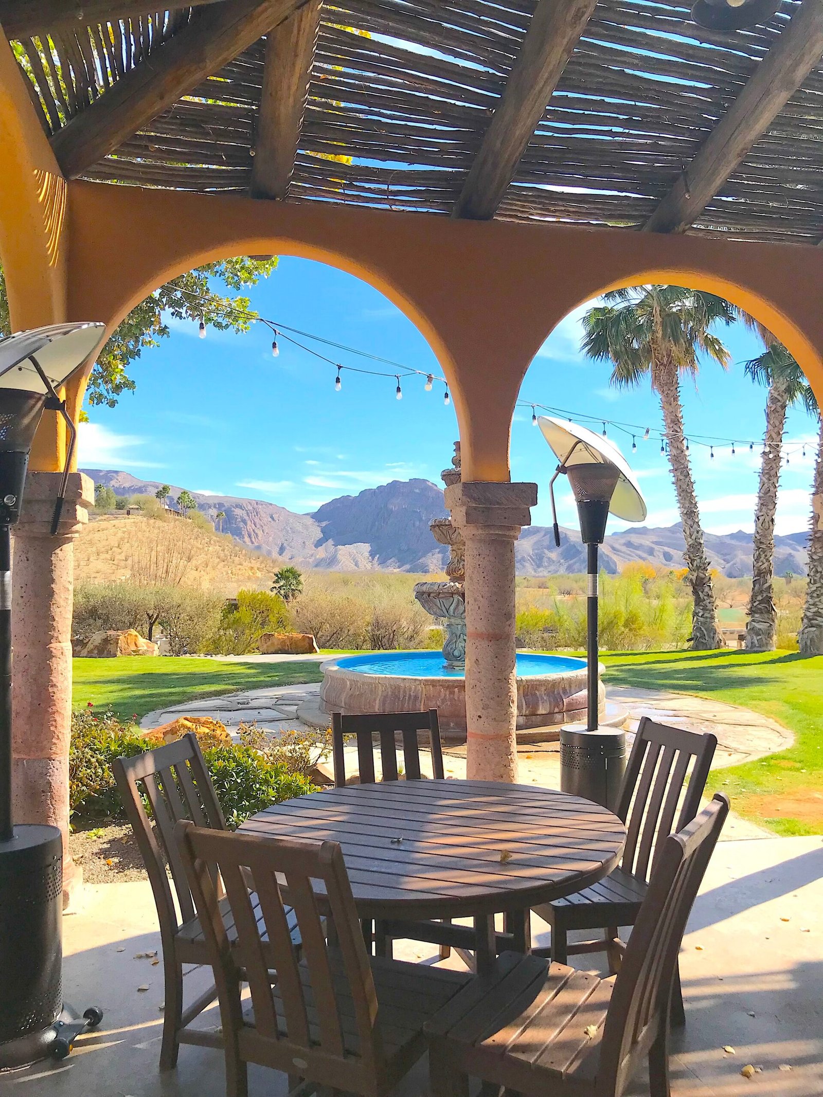 Shaded patio area at Lajitas Resort with Spanish-style archways, wooden tables, outdoor heaters, and a central fountain looking out toward desert palm trees and mountains.