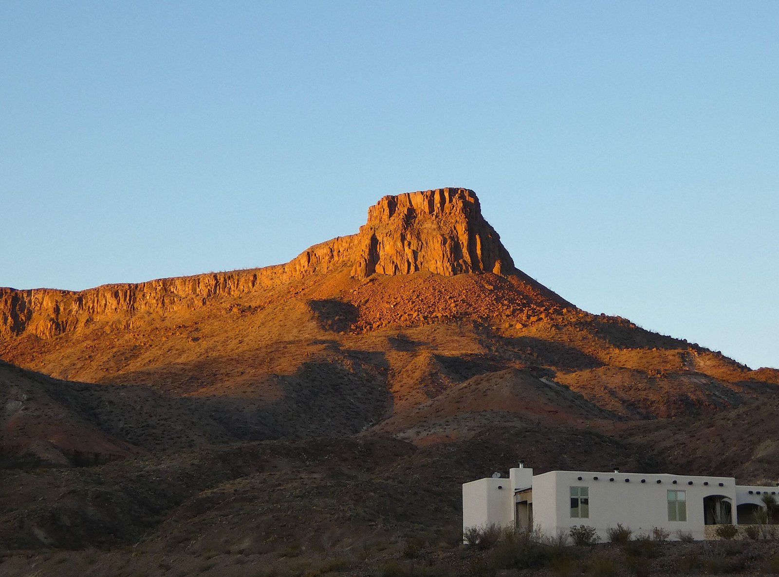 Golden hour sunlight illuminating a high desert mountain peak behind adobe-style resort buildings in Lajitas, Texas.