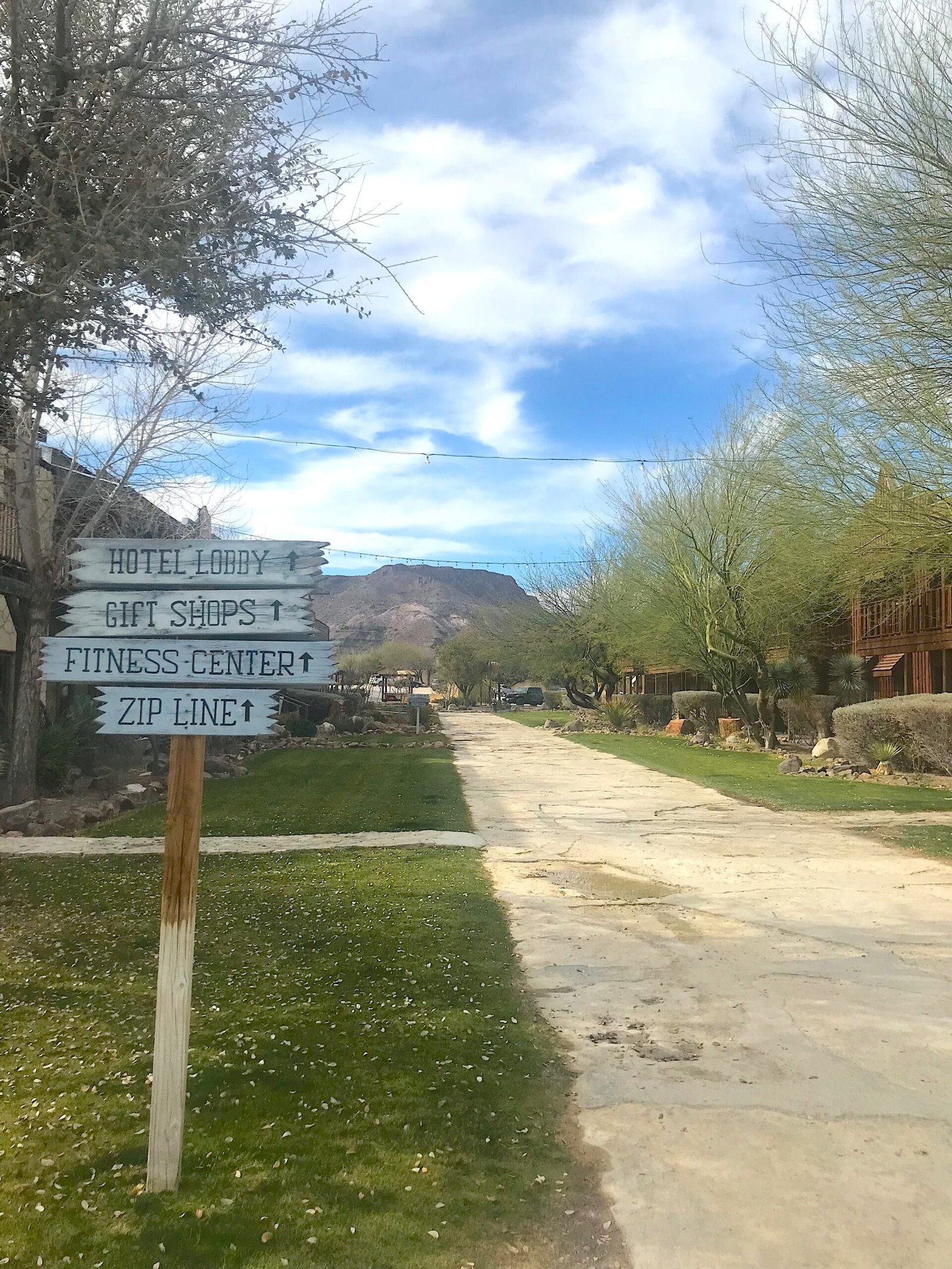 Wooden directional sign on the Lajitas Resort boardwalk pointing toward the zipline, fitness center, gift shops, and hotel lobby.