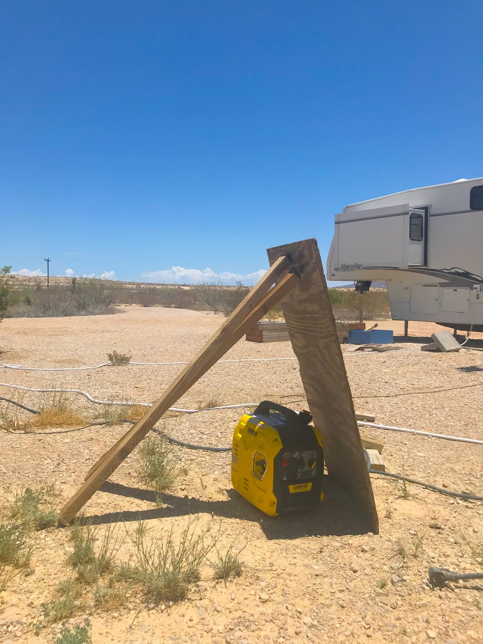 Portable generator set up in the desert gravel with a wooden board propped over it for sun protection during a power outage.