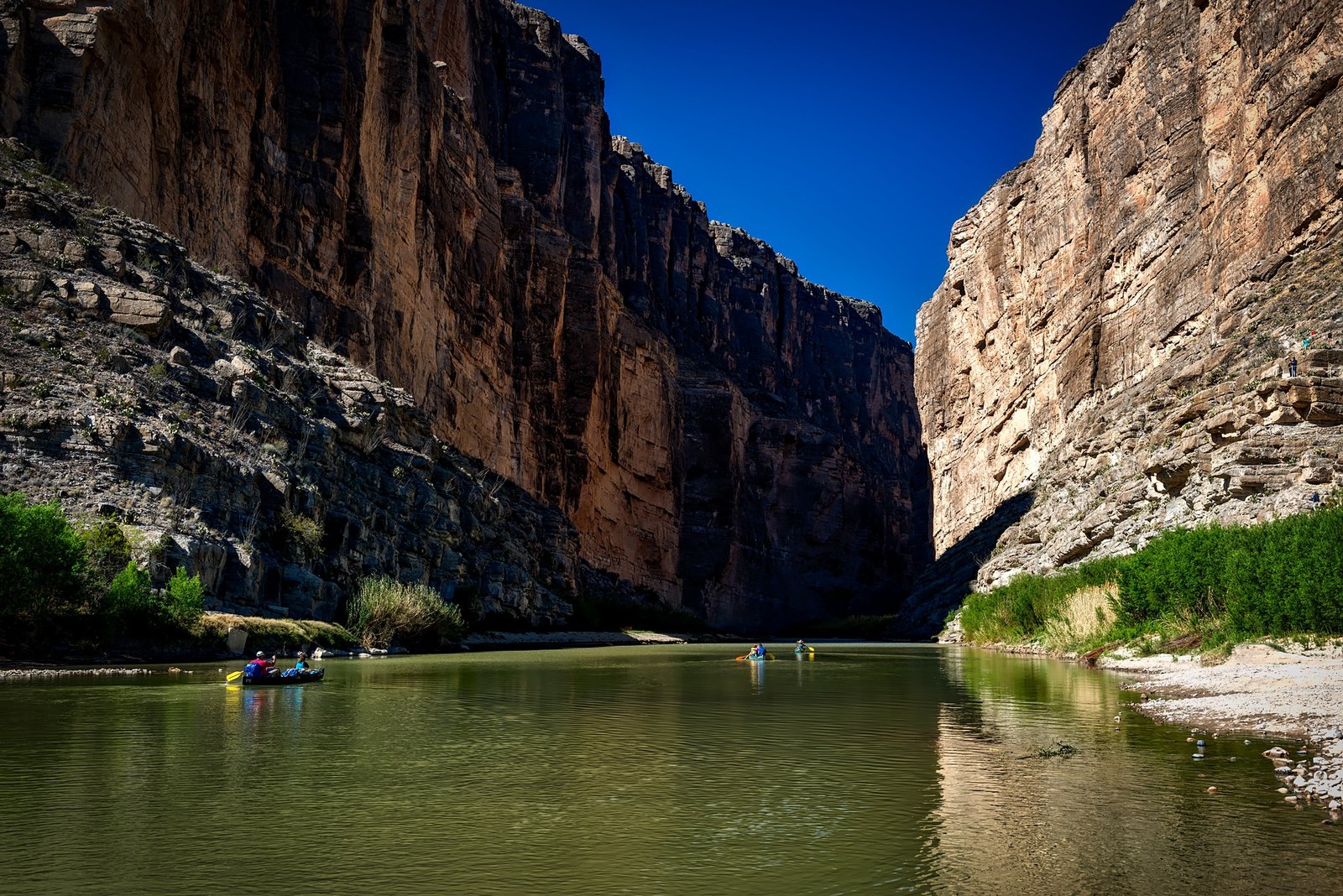 Kayakers paddling on the calm Rio Grande river surrounded by towering vertical rock cliffs in Big Bend Ranch State Park.