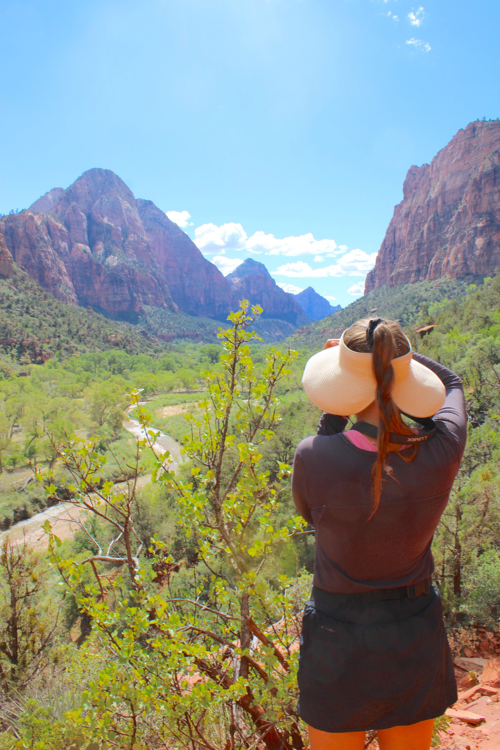A hiker wearing a sun hat looking out at the sweeping green valley and towering sandstone cliffs from the elevated Kayenta Trail in Zion National Park.