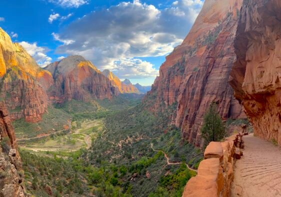 Panoramic landscape view of Zion Canyon with dramatic red rock cliffs, lush green valley, and a winding trail under a cloudy sky in Zion National Park