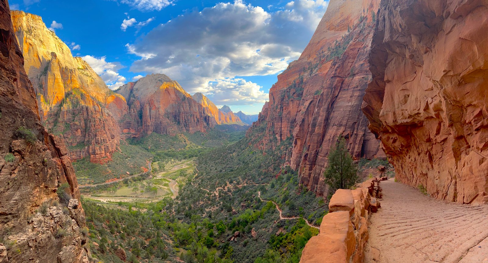 Panoramic landscape view of Zion Canyon with dramatic red rock cliffs, lush green valley, and a winding trail under a cloudy sky in Zion National Park