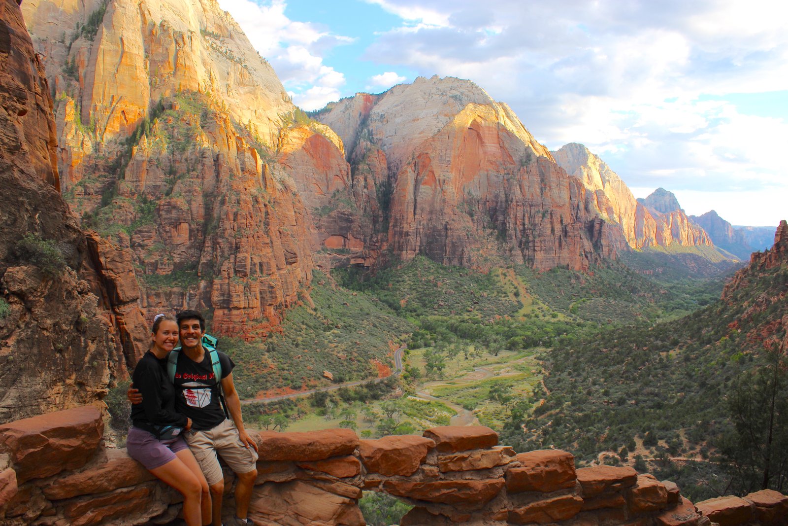 A smiling couple sitting together and looking out over the sprawling Zion Canyon landscape from an elevated viewpoint along the West Rim Trail.