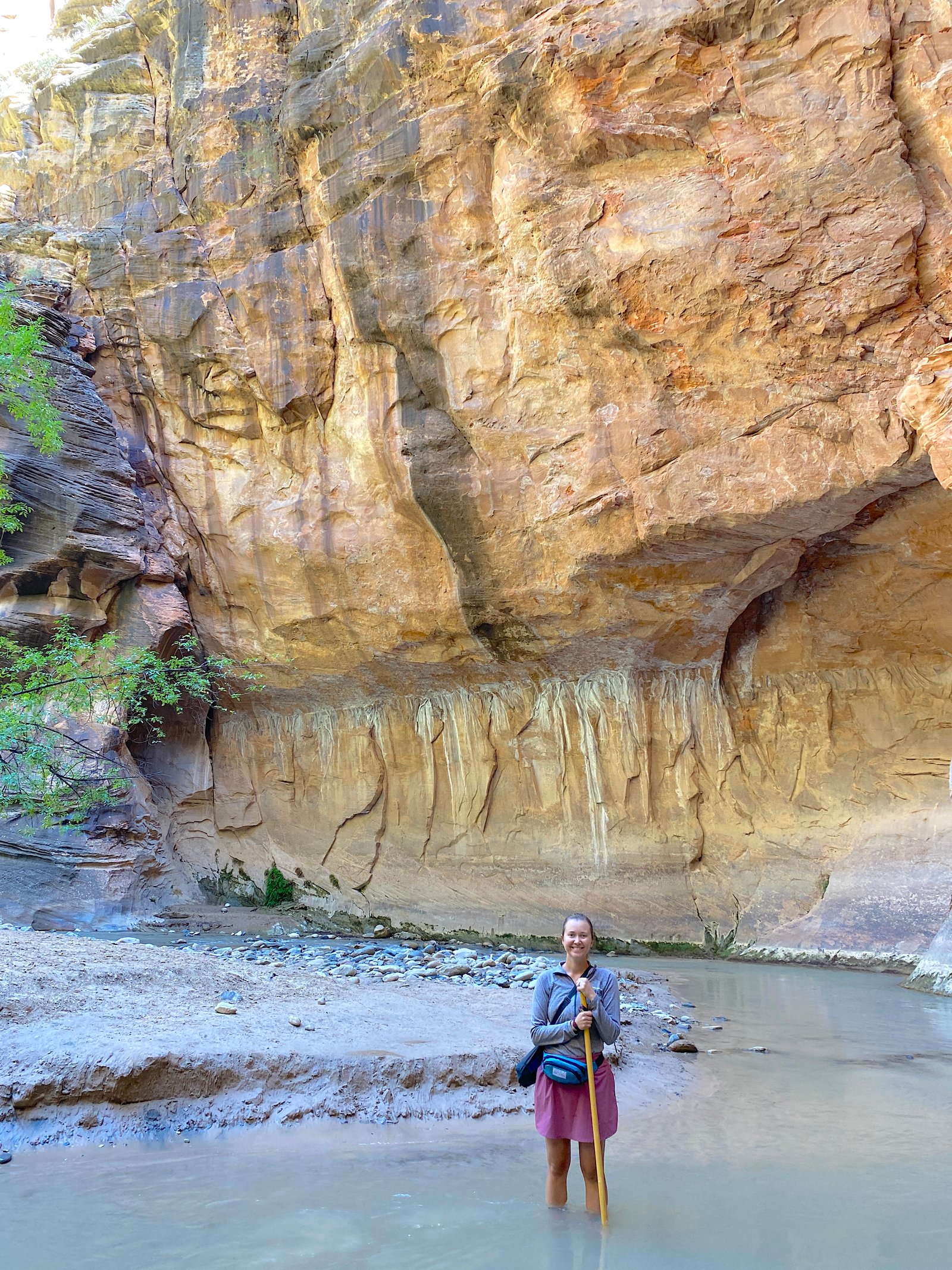 A smiling female hiker standing calf-deep in the flowing river surrounded by towering canyon walls in The Narrows at Zion National Park.