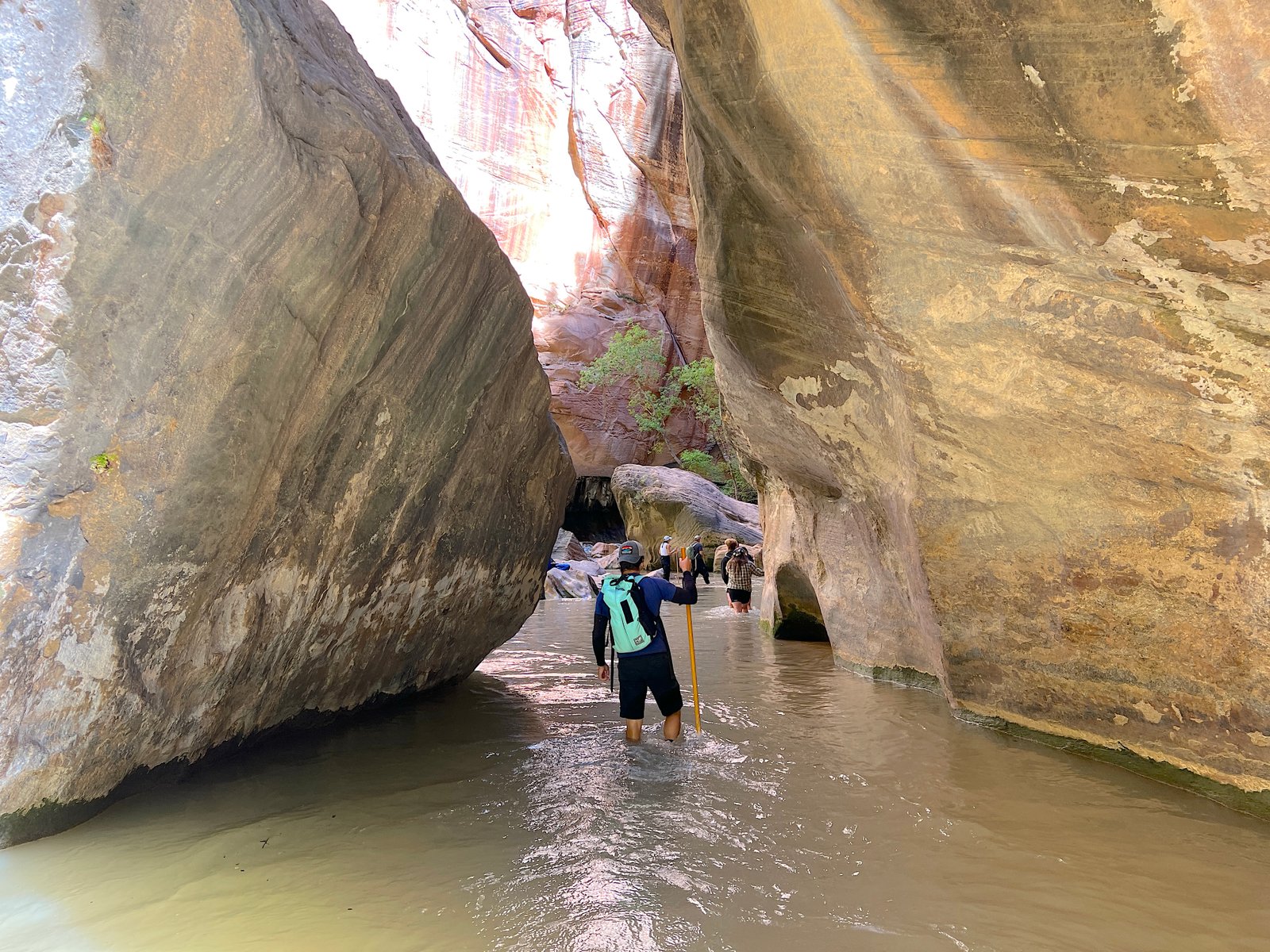 A hiker wading through calf-deep river water between a massive boulder and a towering canyon wall in The Narrows at Zion National Park.