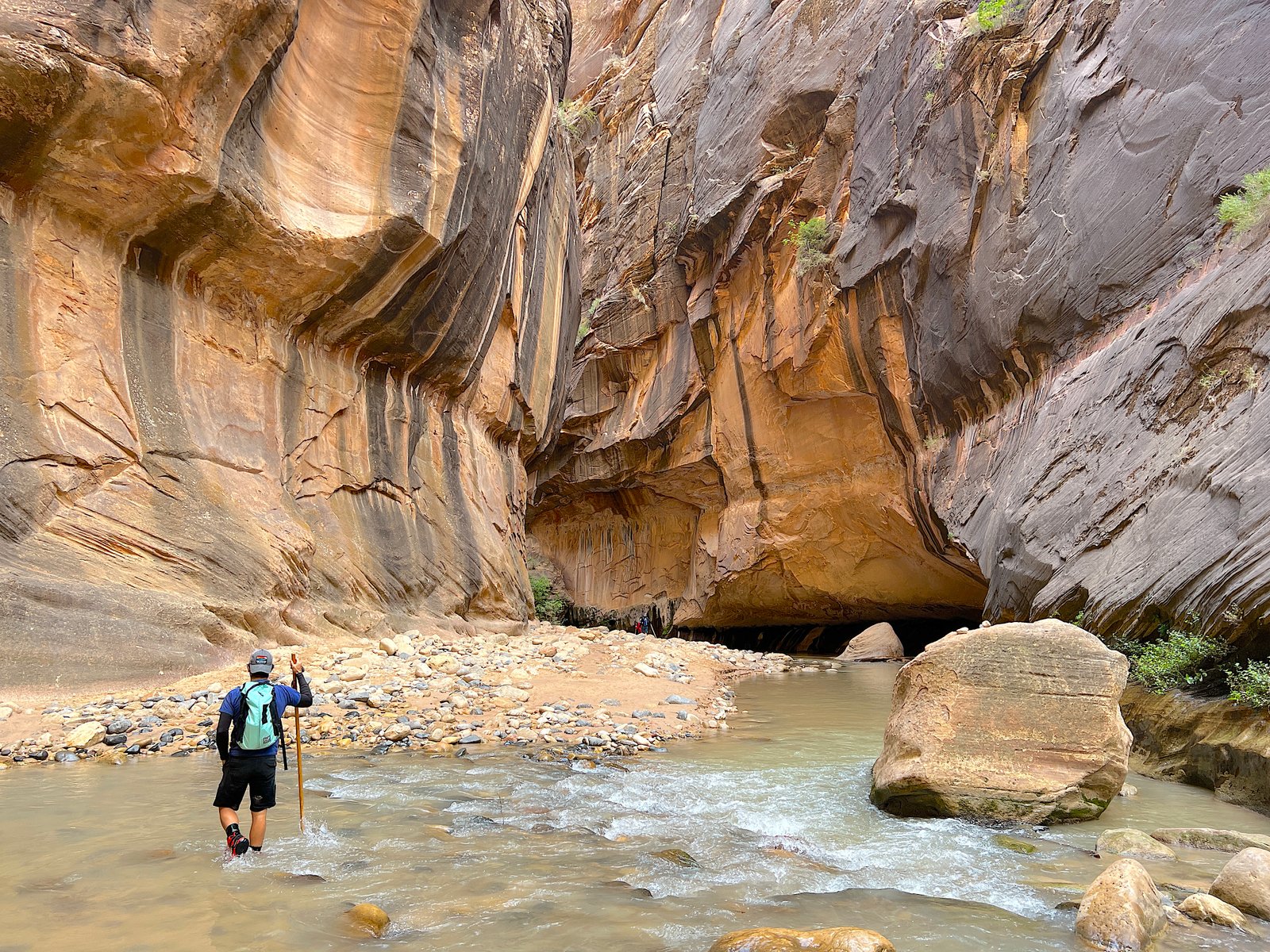 Hiker navigating the rocky riverbed of The Narrows under towering sandstone walls in Zion National Park.
