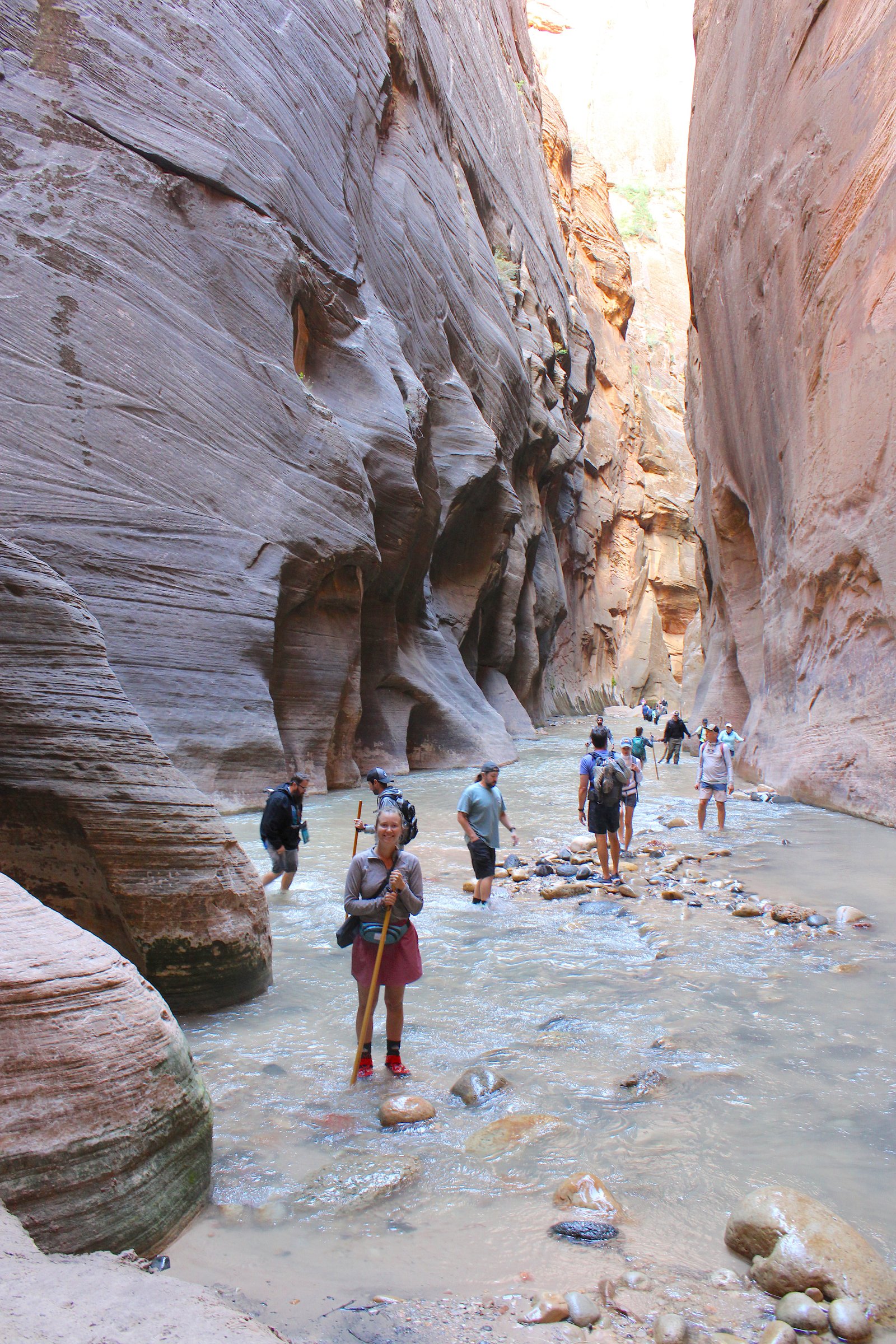A smiling hiker standing beneath towering, close canyon walls with other visitors visible in the background of The Narrows in Zion National Park.
