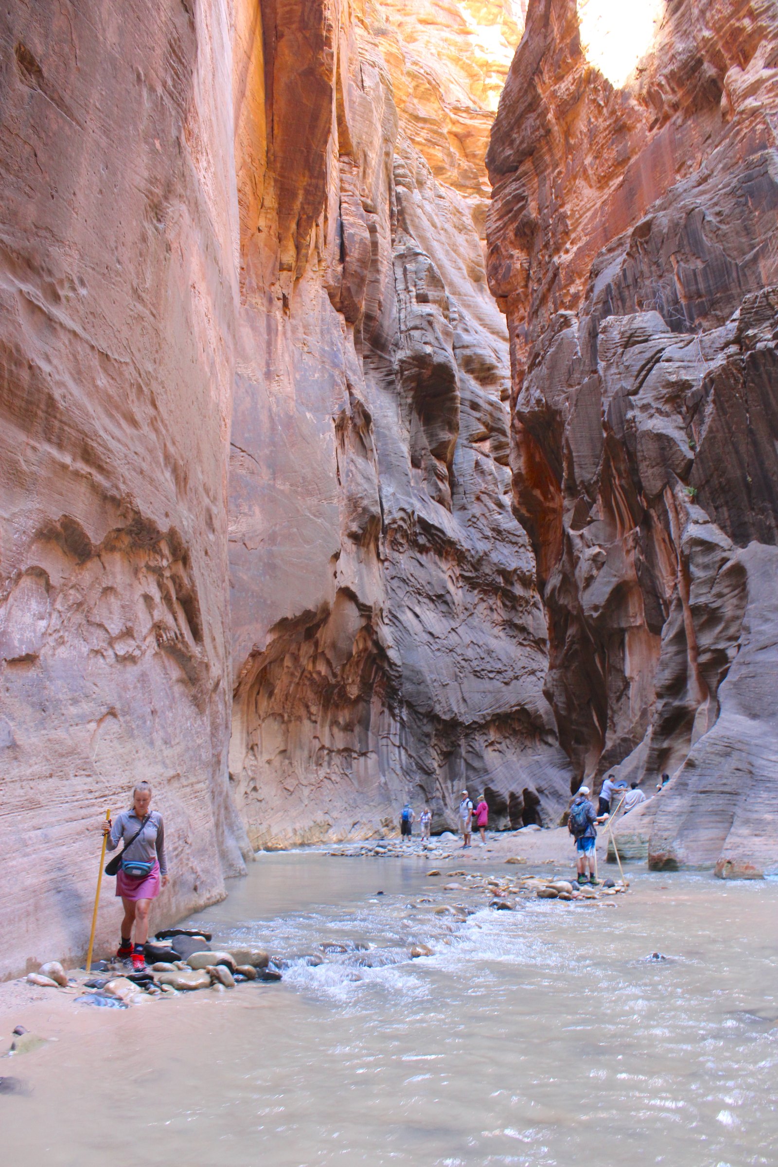 A lone hiker in the distance navigating the winding river beneath the towering red rock walls of The Narrows in Zion National Park.