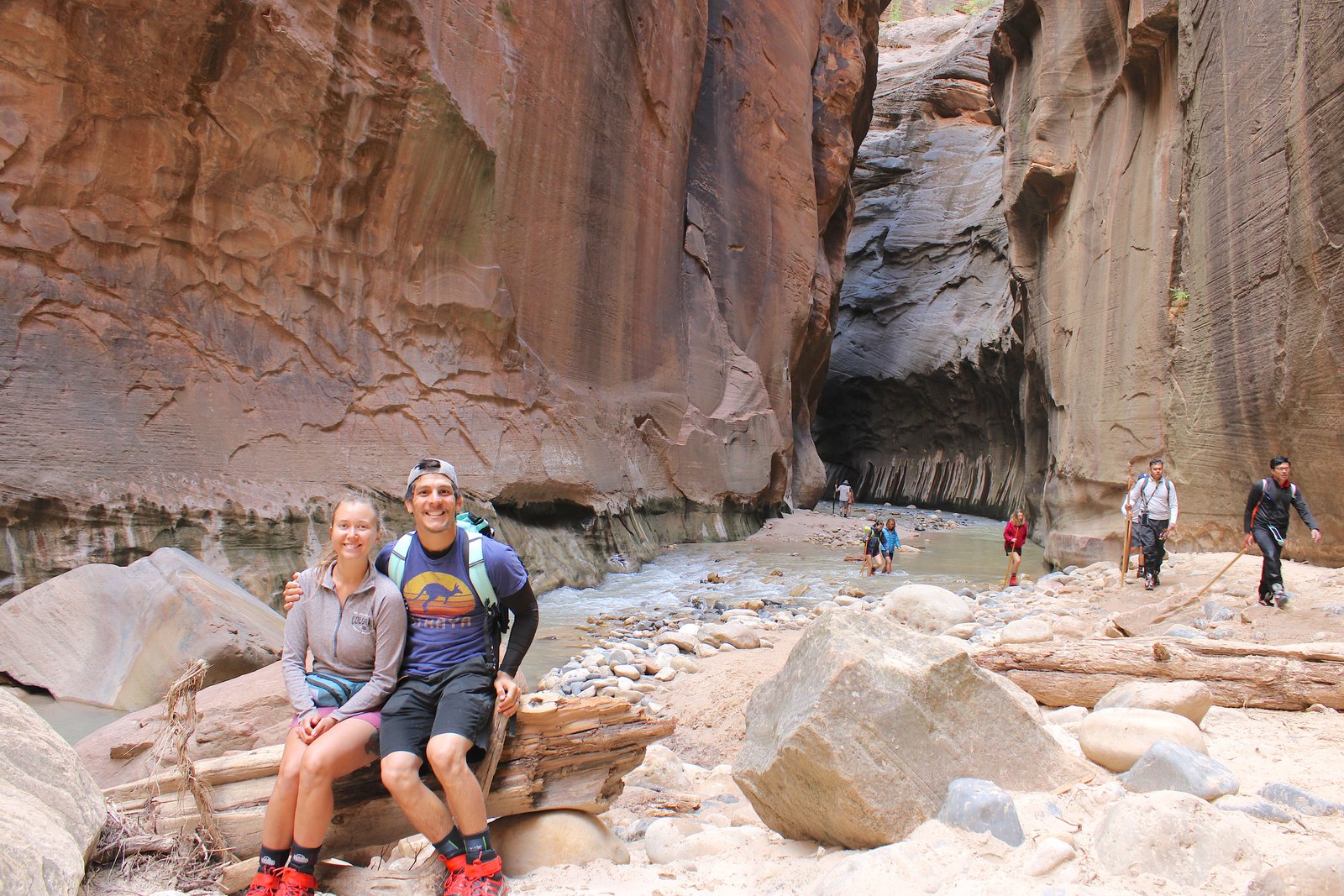 A smiling couple sitting together on a large boulder with the towering stone walls of The Narrows rising up behind them in Zion National Park.