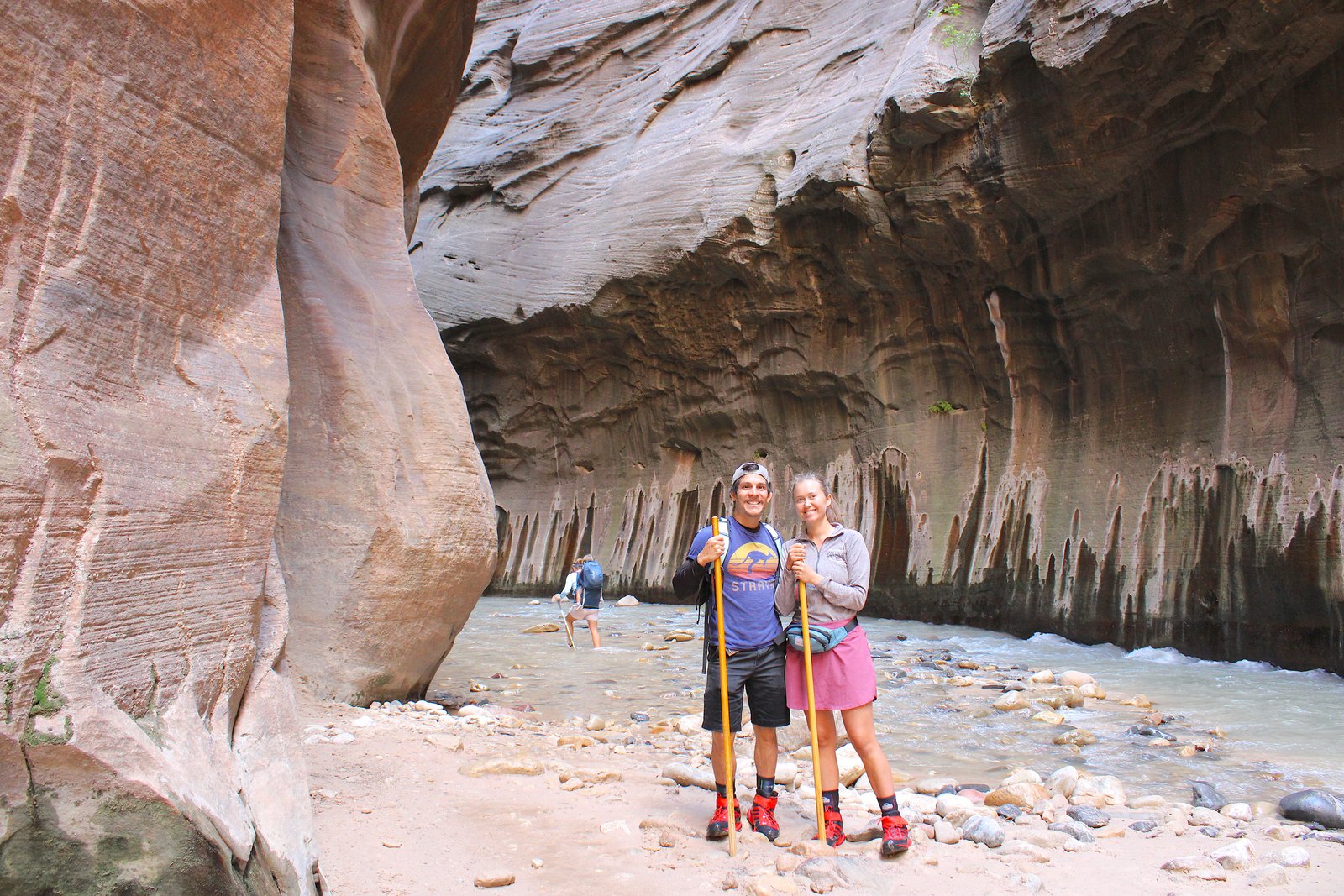 A smiling couple standing together in full gear inside the towering canyon of The Narrows in Zion National Park.