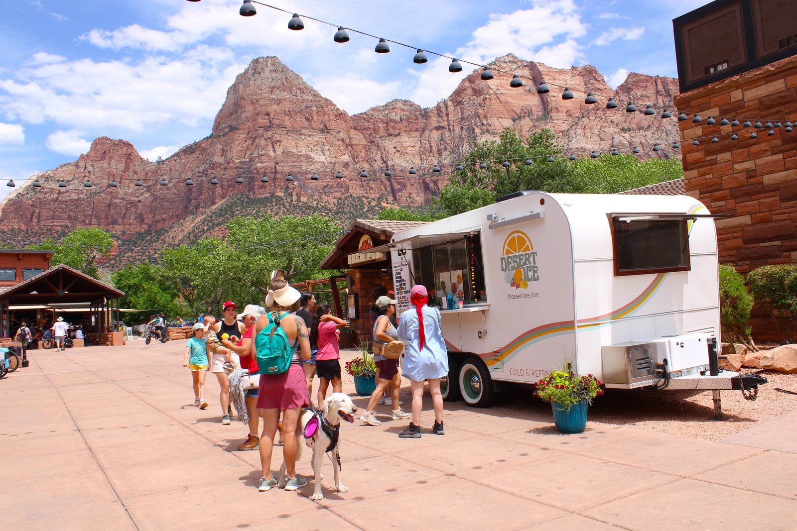 People gathered around the colorful Desert Ice food truck in Zion Canyon Village in Springdale, Utah, with red rock canyon walls rising in the background.