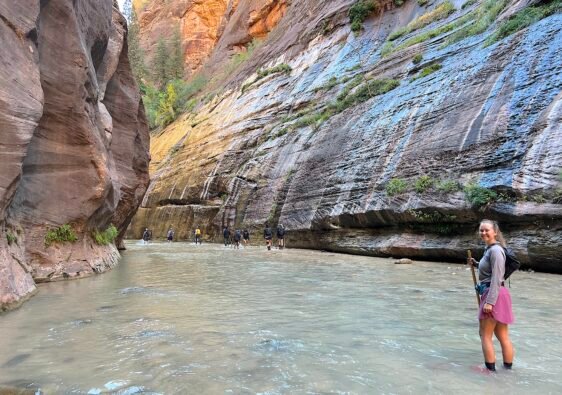 A smiling hiker standing in the flowing Virgin River with a wooden walking stick during the bottom-up hike through The Narrows in Zion National Park.