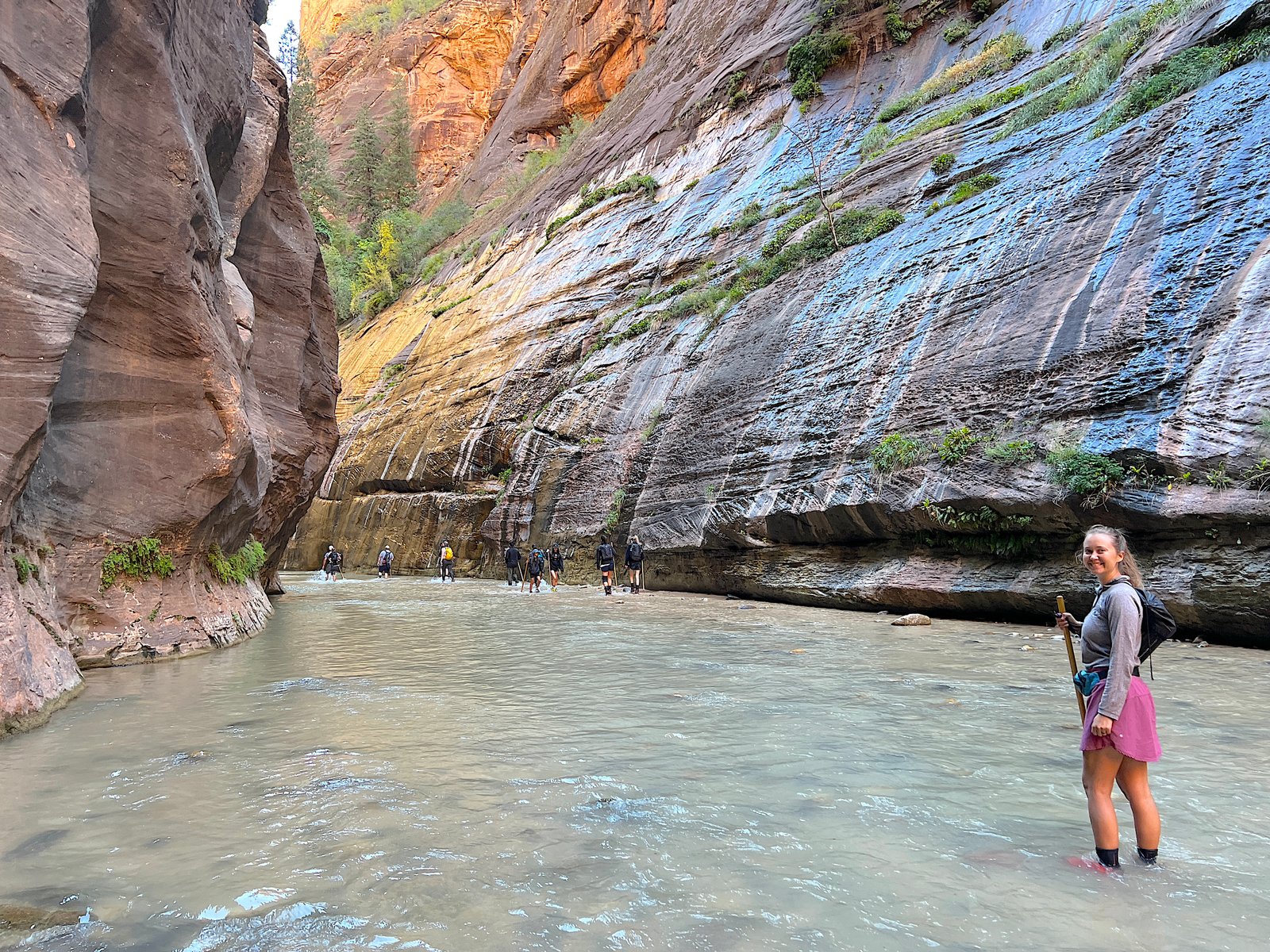 A smiling hiker standing in the flowing Virgin River with a wooden walking stick during the bottom-up hike through The Narrows in Zion National Park.