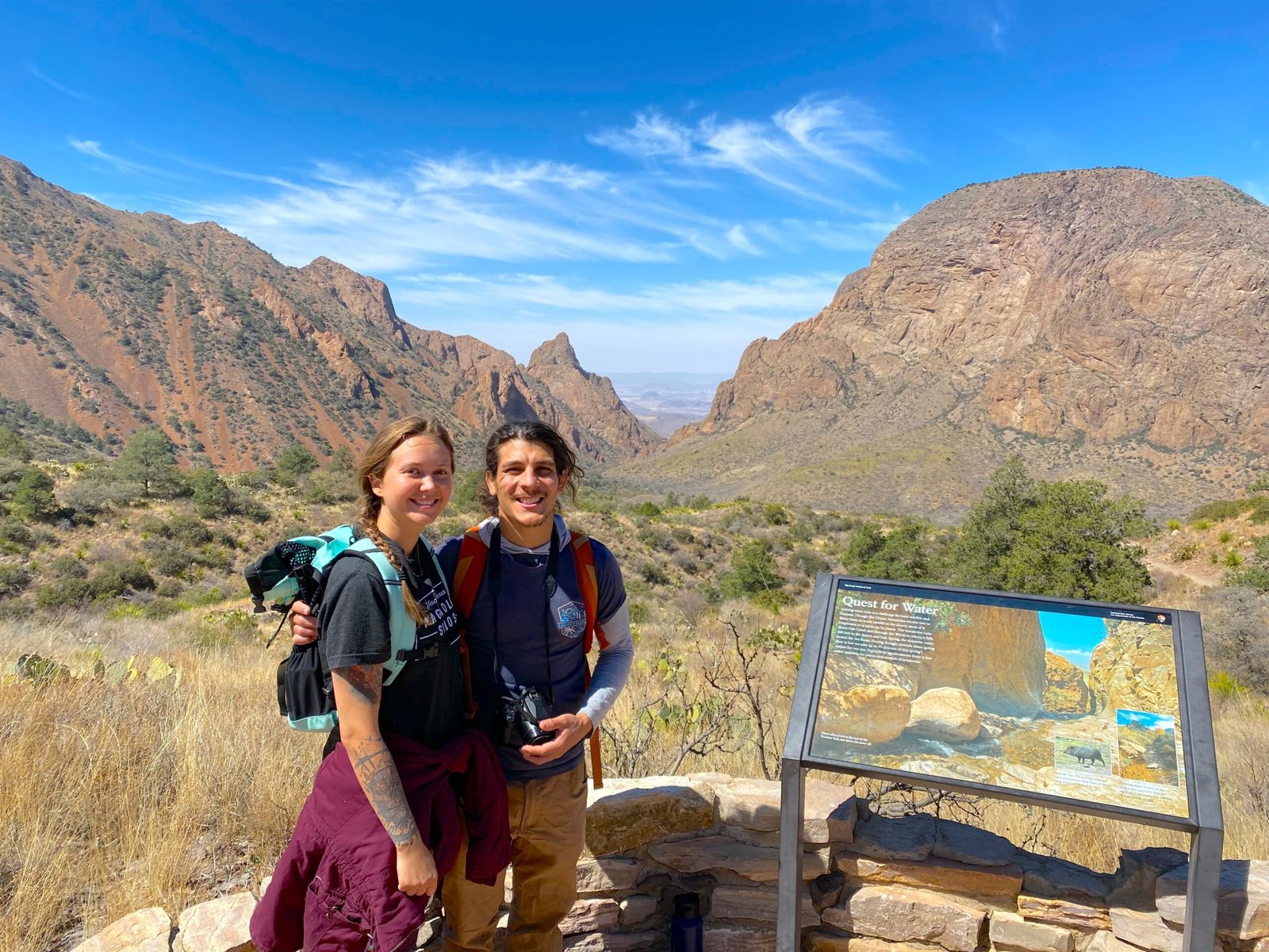 Two hikers smiling in front of an informational trail map sign in a desert mountain park.