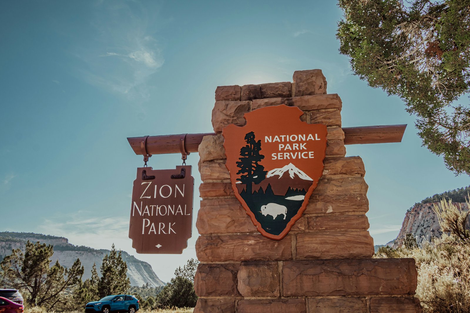 The stone entrance sign for Zion National Park under a bright blue sky with surrounding desert landscape and trees.