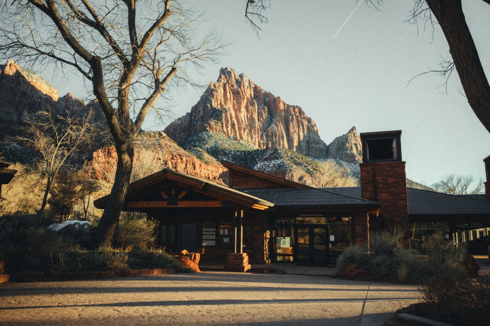 The Zion National Park Visitor Center building with the massive peak of Watchman Mountain rising dramatically behind it under a blue sky.