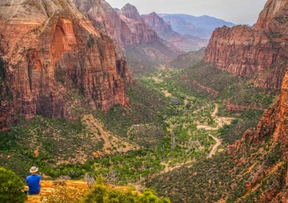 Panoramic view of Zion Canyon with towering red sandstone cliffs, lush green valley floor, and a winding river from an overlook trail in Utah.