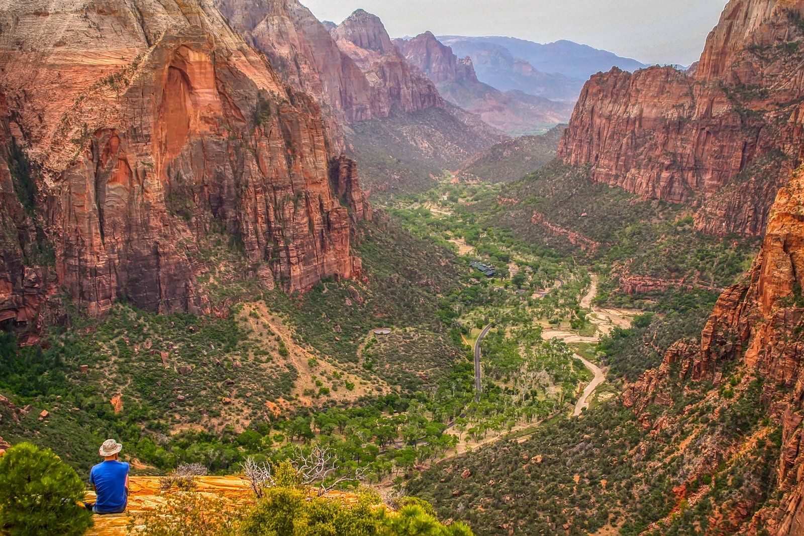 Panoramic view of Zion Canyon with towering red sandstone cliffs, lush green valley floor, and a winding river from an overlook trail in Utah.