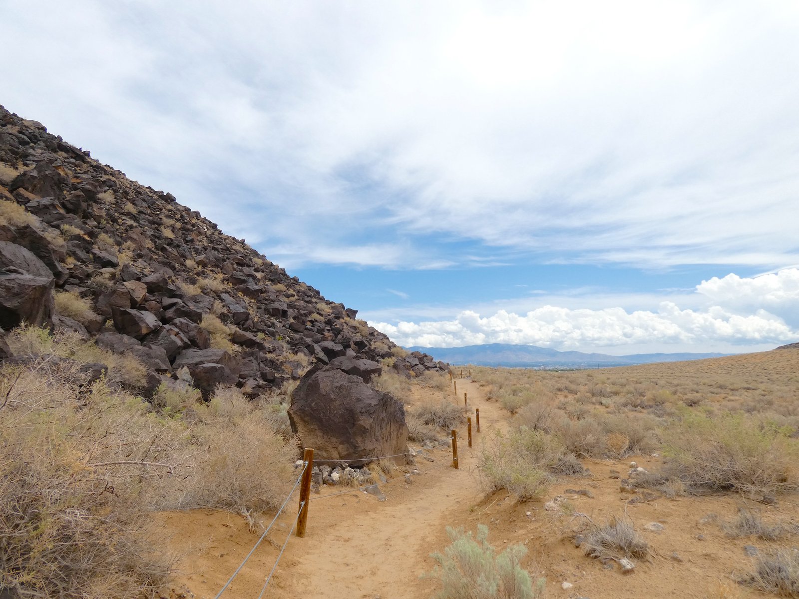 A sandy hiking trail winding alongside the towering dark basalt rock wall at Petroglyph National Monument.