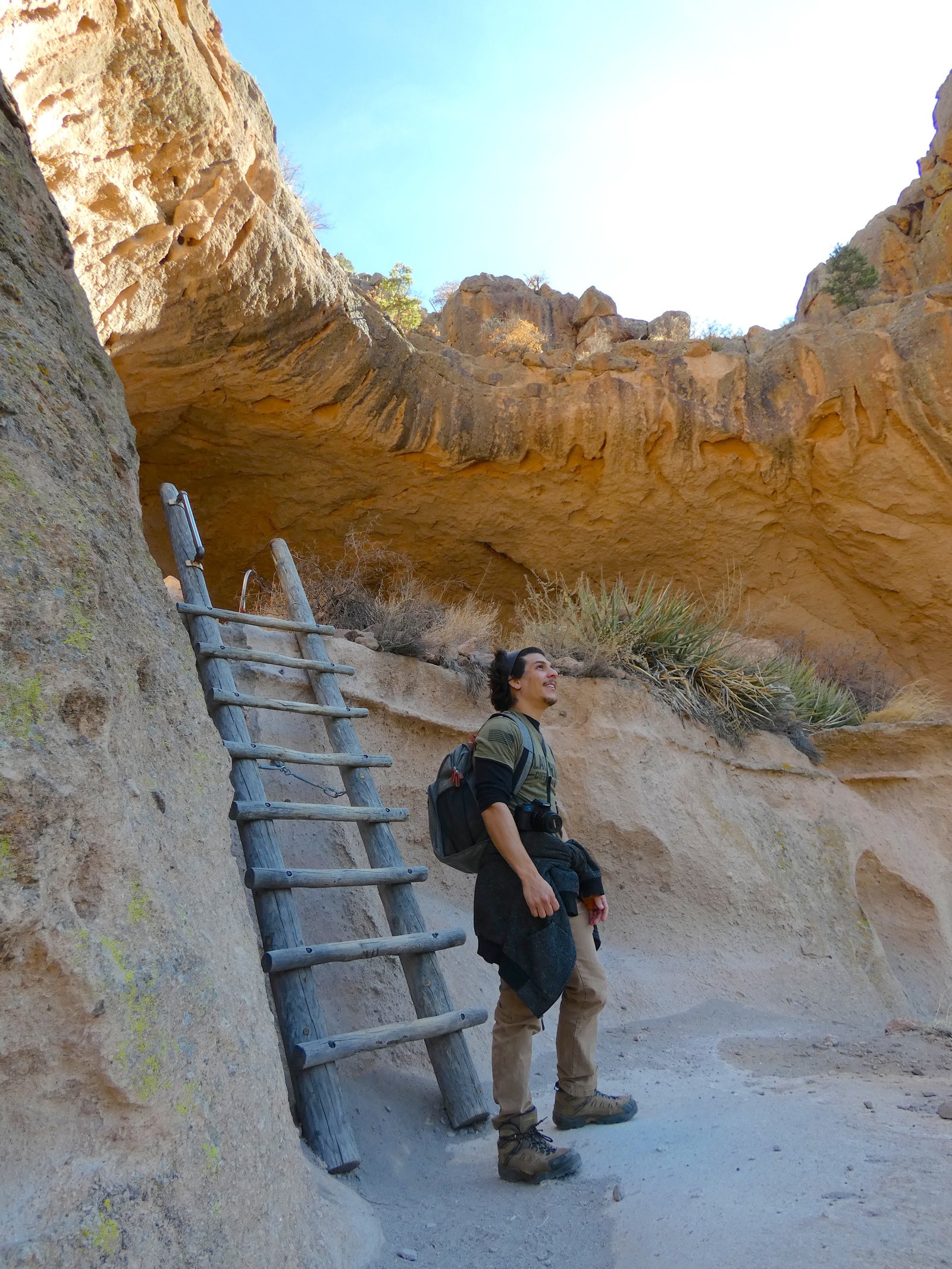 A hiker wearing a backpack standing next to the wooden ladder at the base of Alcove House in Bandelier National Monument.